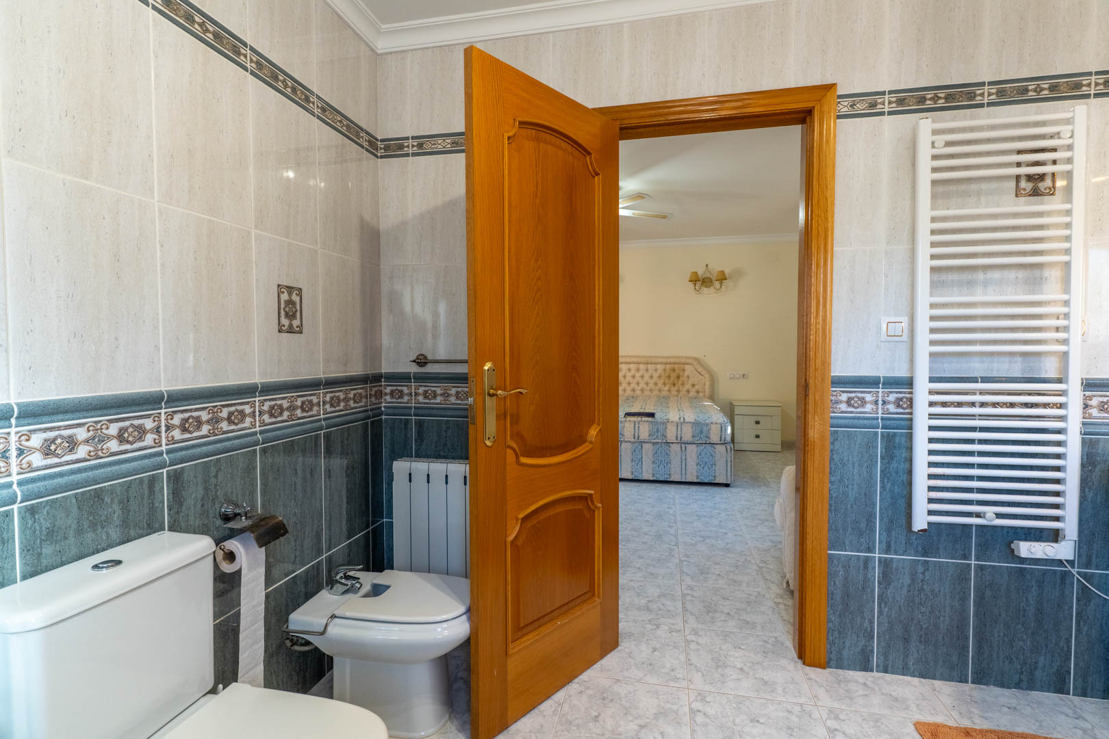 Zicht vanuit de en-suite badkamer op de slaapkamer, voorzien van tweekleurige betegeling, een witte handdoekradiator en een massief houten deur.