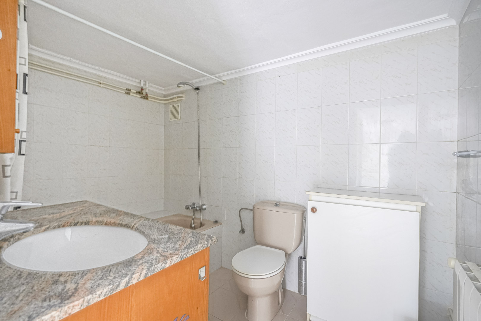 Badkamer met granieten werkblad, inbouwwastafel, beige toilet en wit betegelde wanden. Voorzien van een douchegedeelte en een wandradiator.