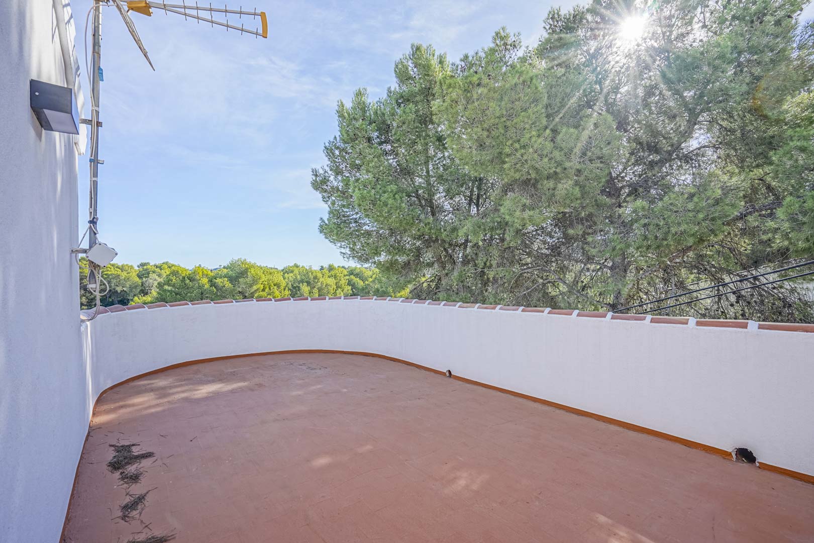 Bovenliggend terras met terracotta tegels en een gebogen witte borstwering, uitkijkend over de groene pijnbomen in een zonnige omgeving.