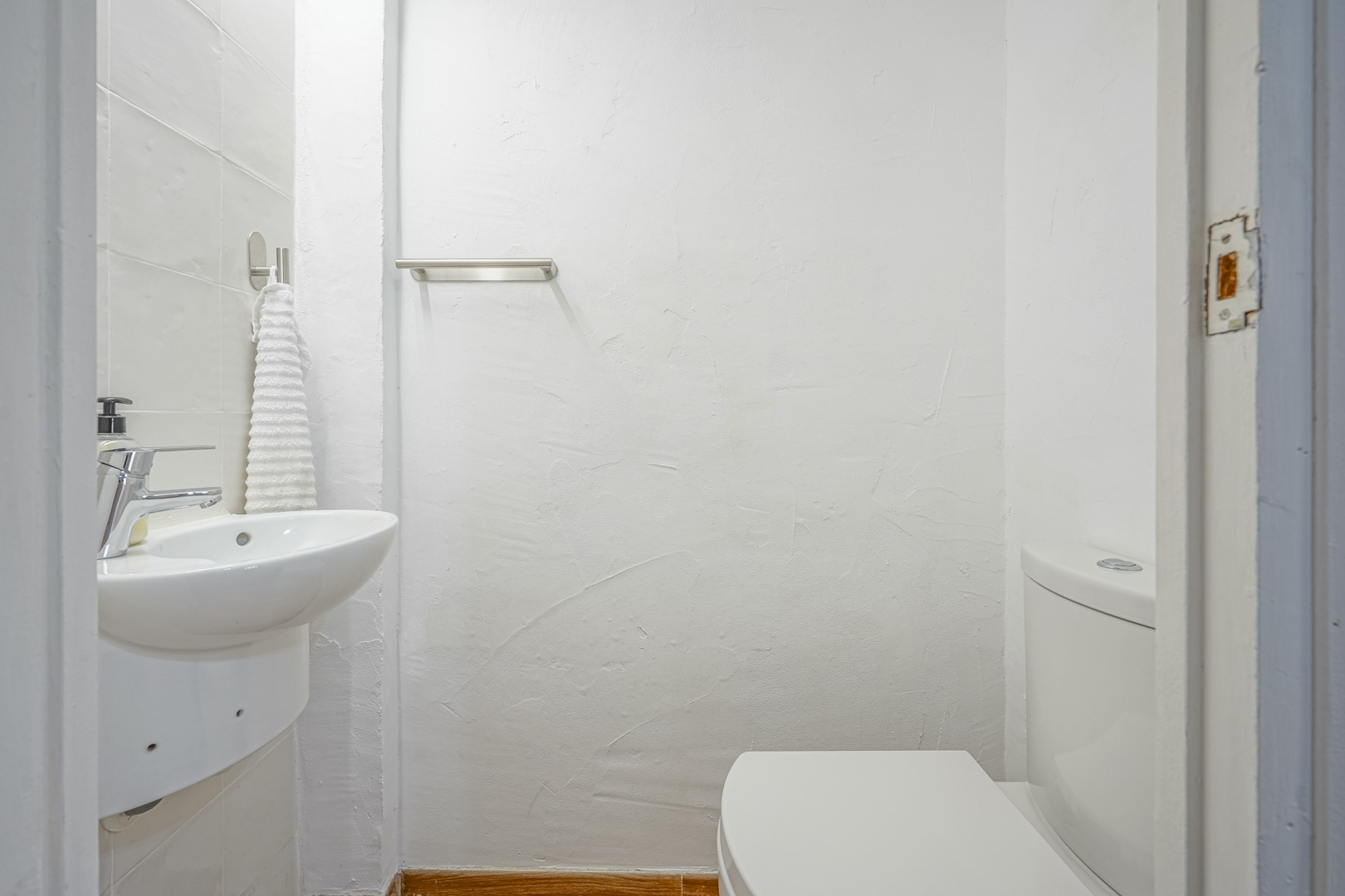 Compact gastentoilet met witte structuurwanden, een hangende keramische wastafel met chromen kraan en een roestvrijstalen handdoekrek.