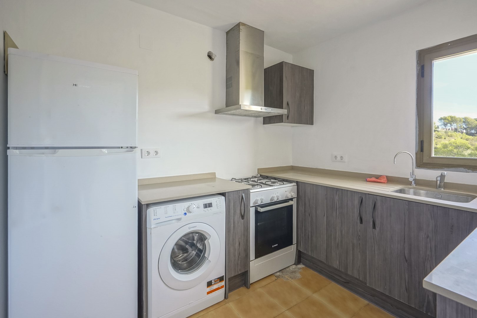 Functionele keuken met grijze kasten in houtoptiek, voorzien van diverse inbouwapparatuur en een raam met uitzicht op de groene omgeving.