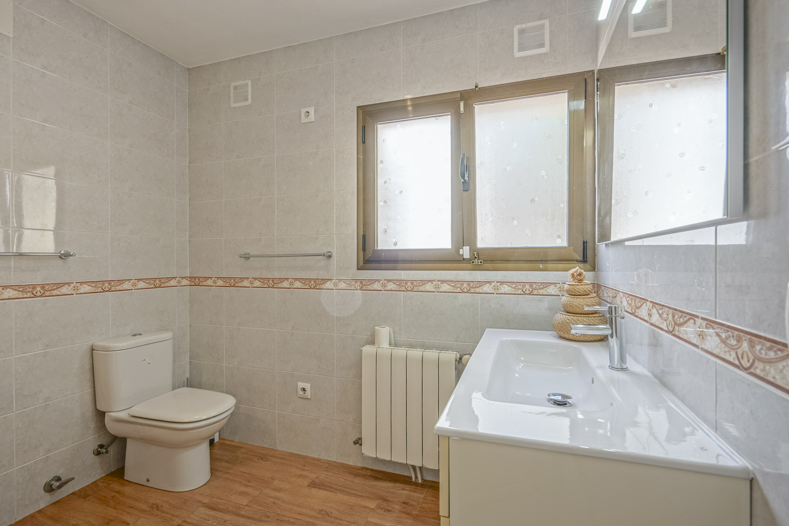 Gerenoveerde badkamer met houtlook vloer, betegelde wanden met decoratieve bies, wit wastafelmeubel en radiator onder een raam met melkglas.