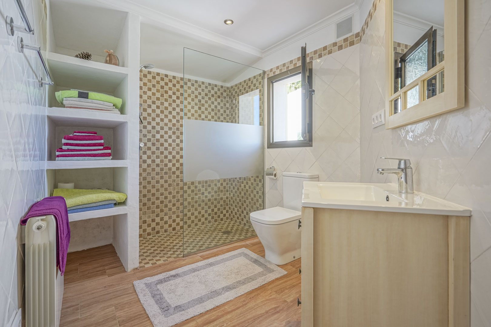 Gerenoveerde badkamer met inloopdouche met mozaïektegels, ingebouwde gemetselde schappen, vloer met hout-look en een wastafelmeubel van licht hout.