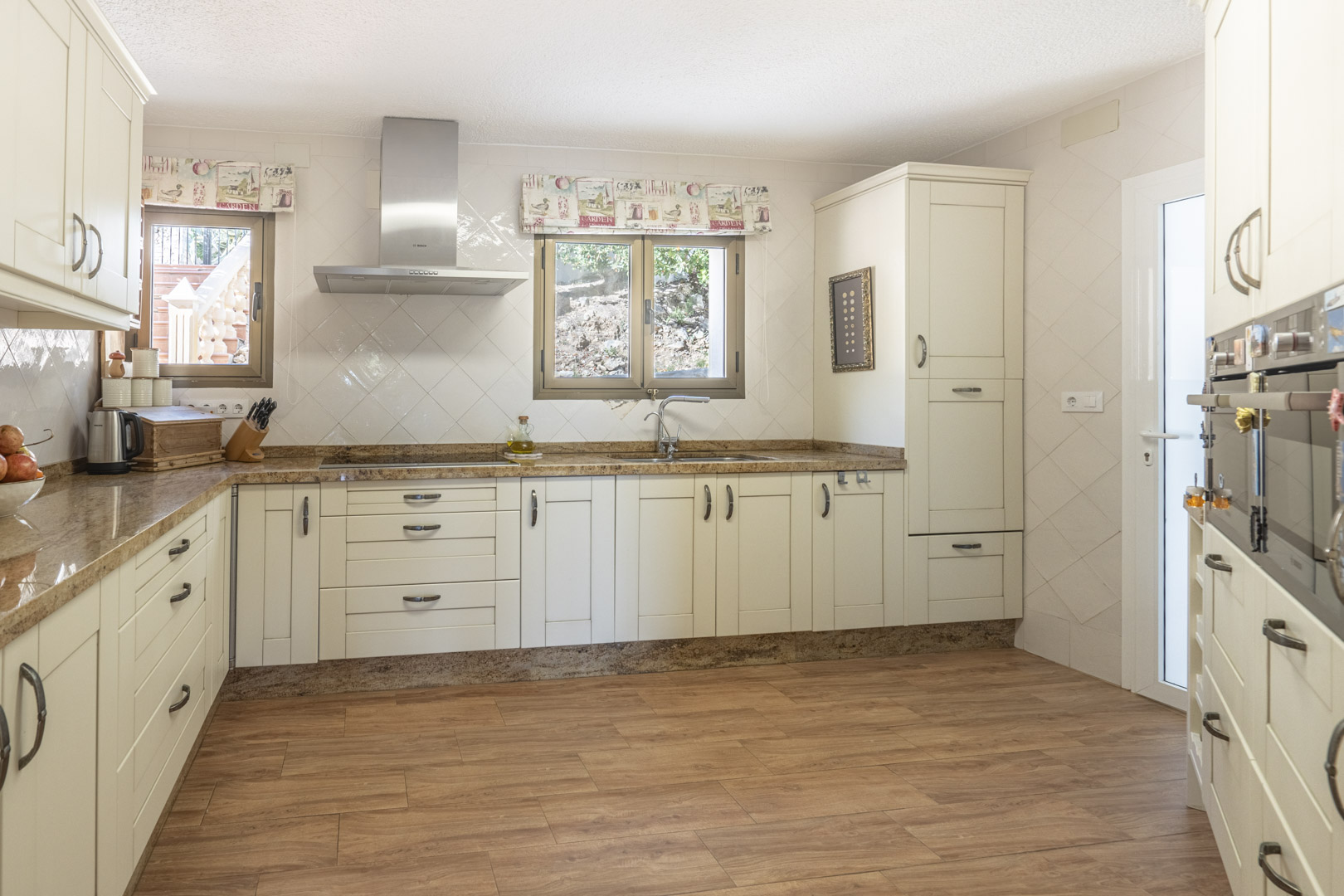 Gerenoveerde keuken met crèmekleurige kasten, granieten werkbladen, rvs afzuigkap en keramische tegels met houtlook. Veel lichtinval door de vensters.