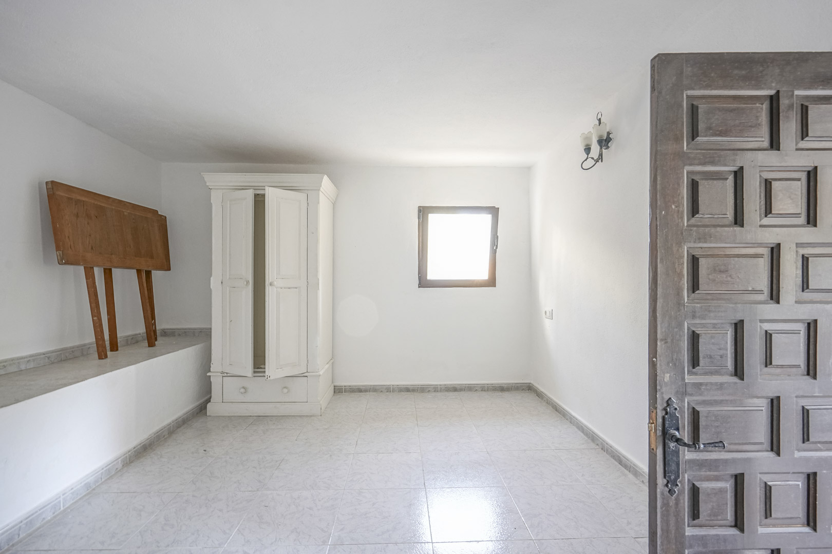 Lichte kamer met een lichte tegelvloer, witte muren en een traditionele bewerkte houten deur. Voorzien van een vrijstaande witte kast en natuurlijke lichtinval.