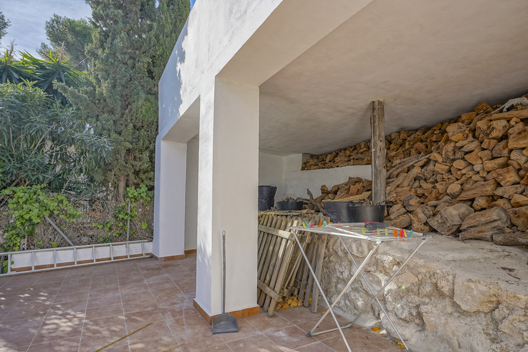 Overdekte buitenopslagruimte met een betegeld terras, witte kolommen en een grote houtopslag tegen een natuurstenen muur.