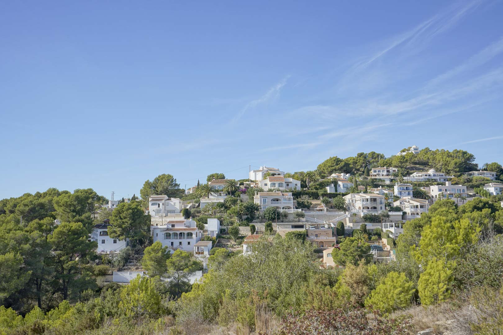 Panoramisch uitzicht op de woonwijk Pinomar in Jávea, met traditionele witte villa's genesteld tegen een beboste heuvel onder een helderblauwe hemel.