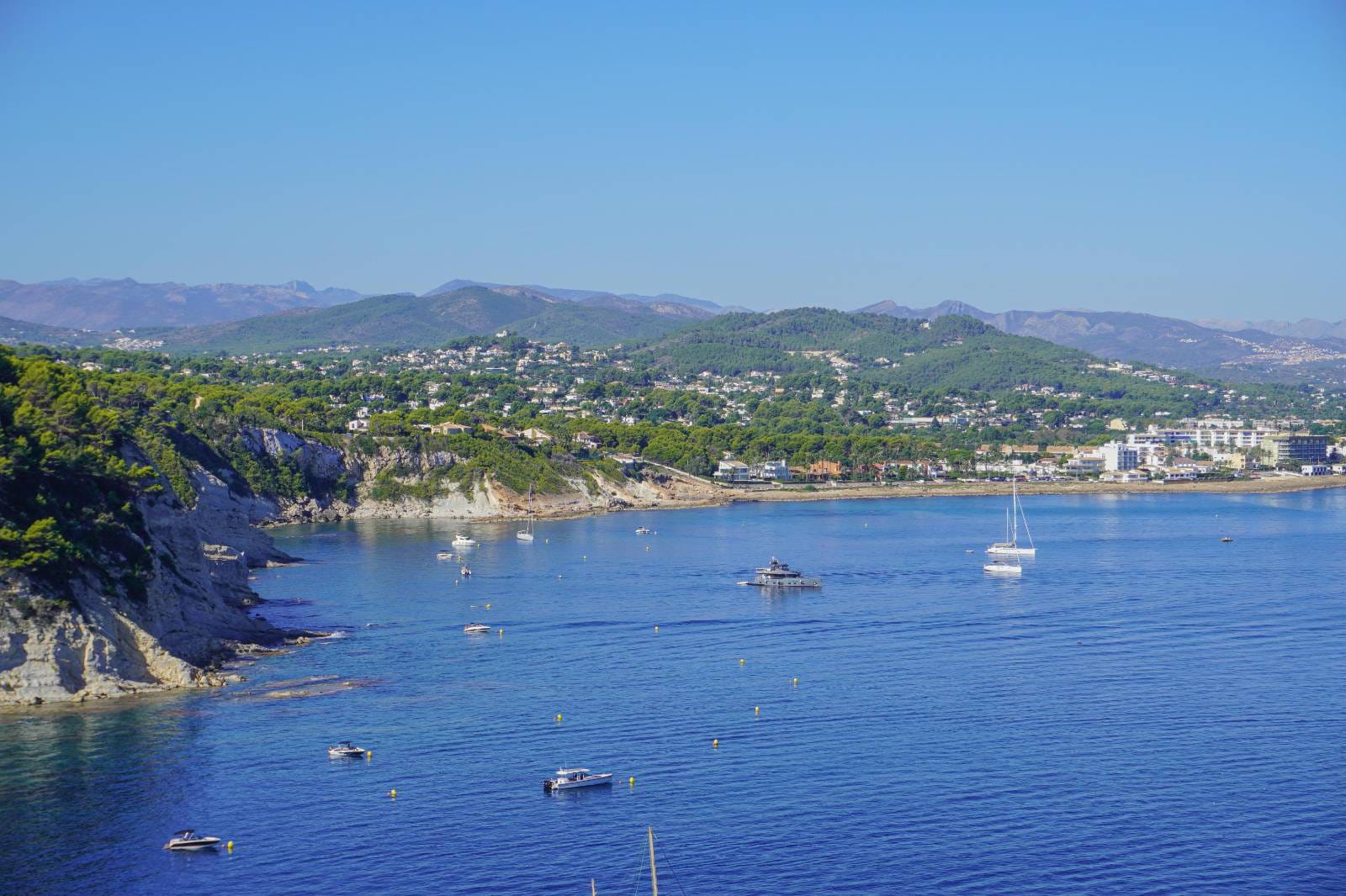 Panoramisch uitzicht over de baai van Jávea met helderblauw water, plezierboten en de rotsachtige kustlijn met dennenbossen en bergen in de verte.