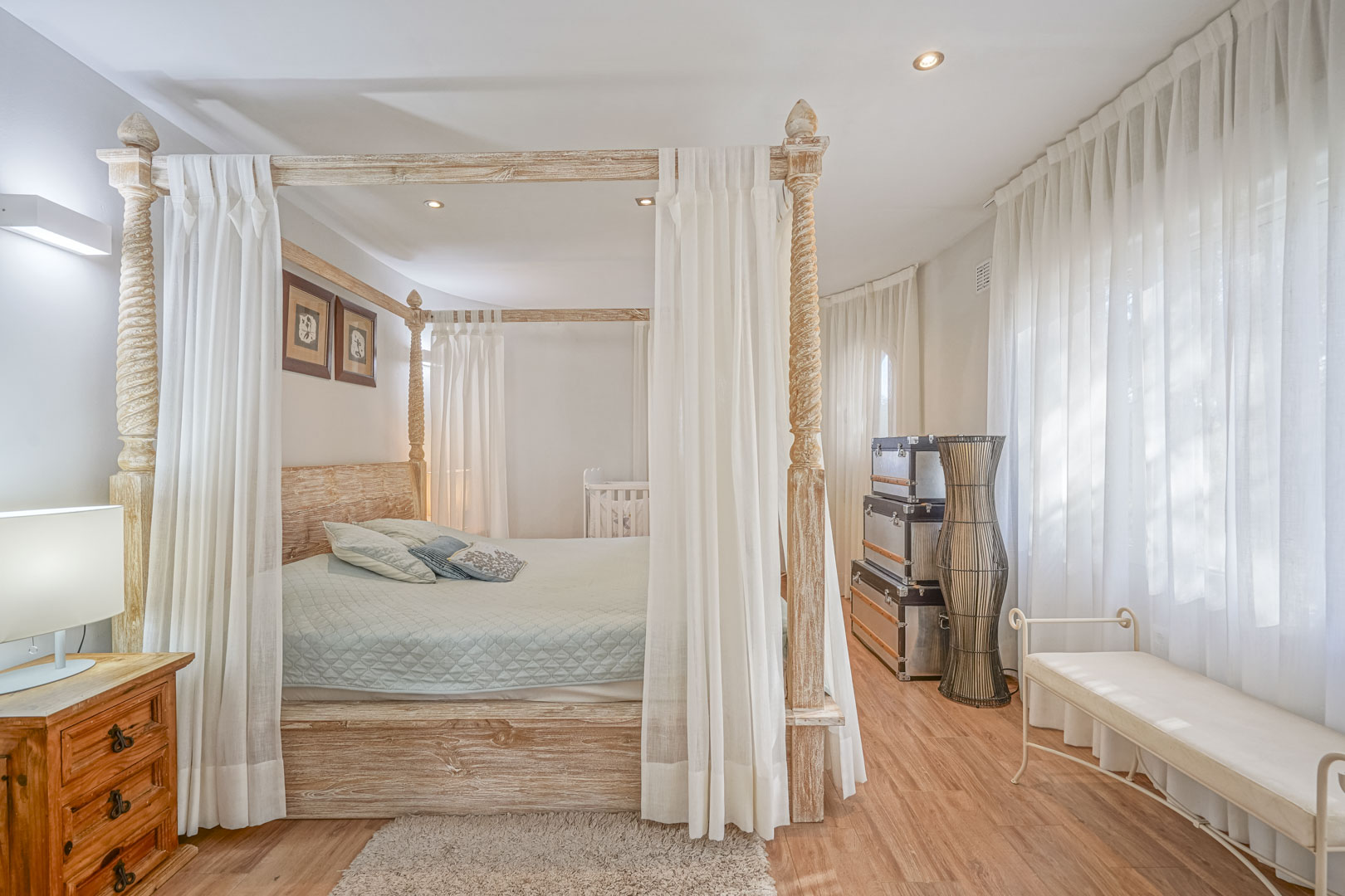 Slaapkamer met een houten hemelbed met houtsnijwerk, laminaatvloer en grote ramen voorzien van witte lichtdoorlatende gordijnen.