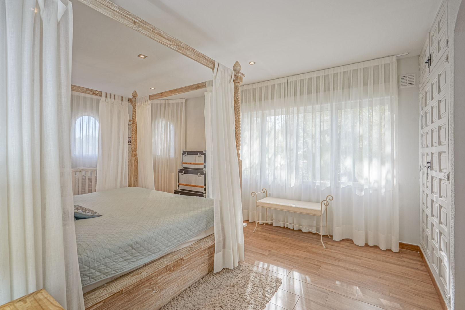 Slaapkamer met een licht houten hemelbed, keramische vloer met houtlook en grote ramen met vitrage die zorgen voor een zachte natuurlijke lichtinval.