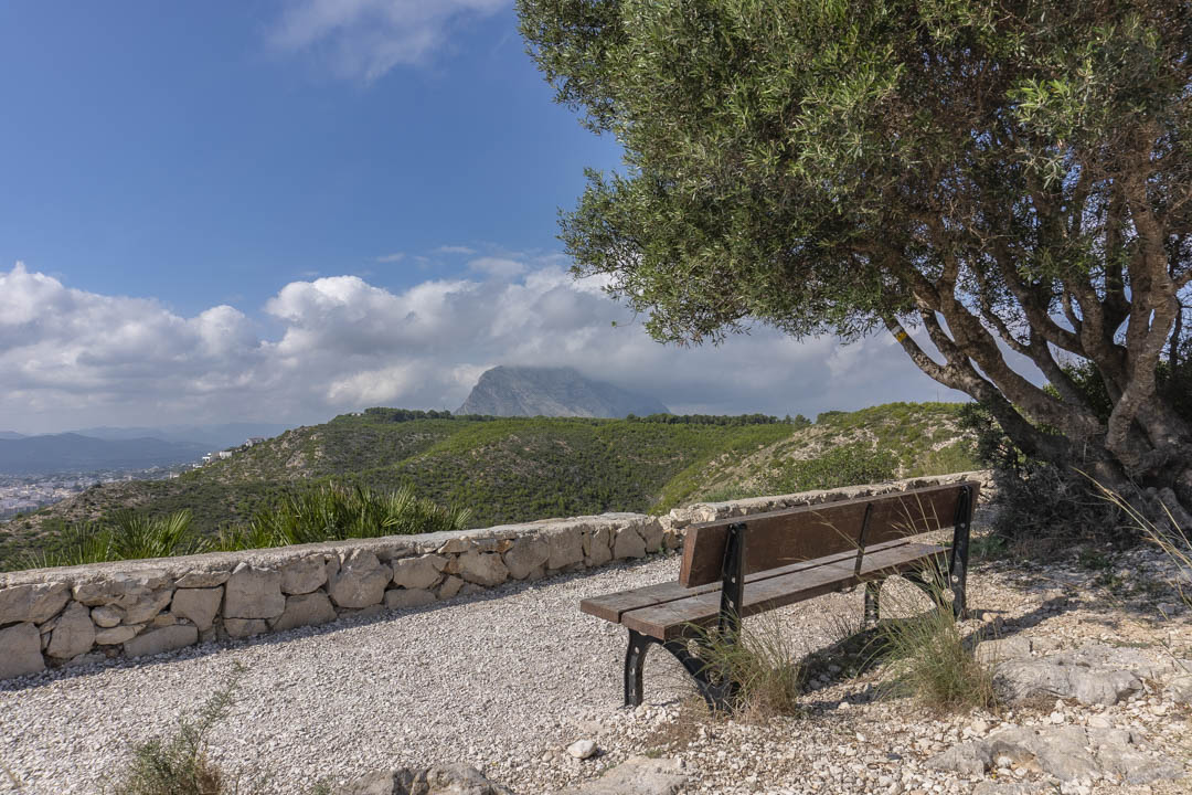 Uitkijkpunt met houten bank en natuurstenen muur, met panoramisch uitzicht op de Montgó-berg en de omliggende groene valleien van Jávea.