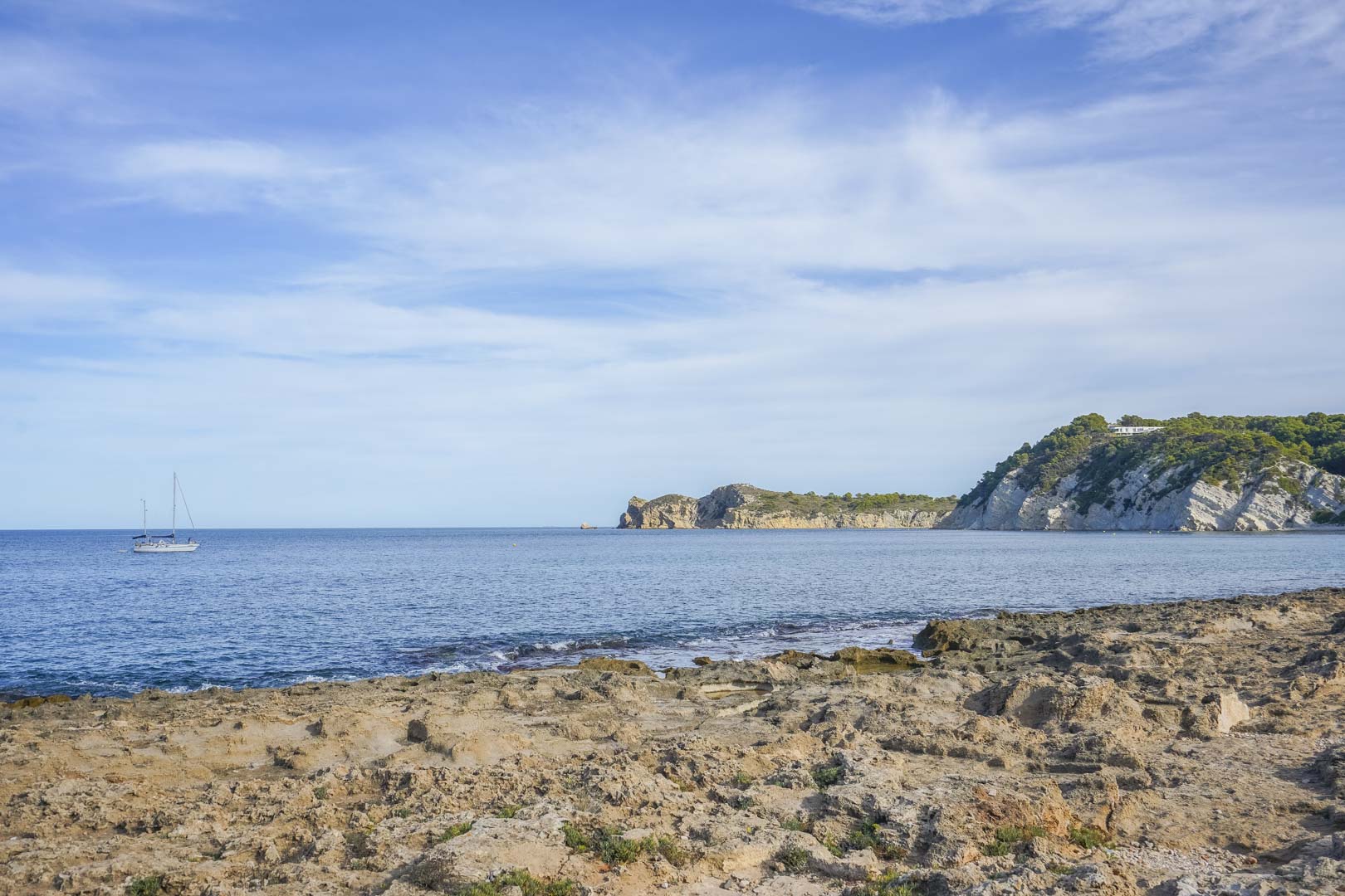 Uitzicht op de rotsachtige kustlijn van Jávea met de Middellandse Zee, kalkstenen kliffen en een zeilboot onder een heldere blauwe lucht.