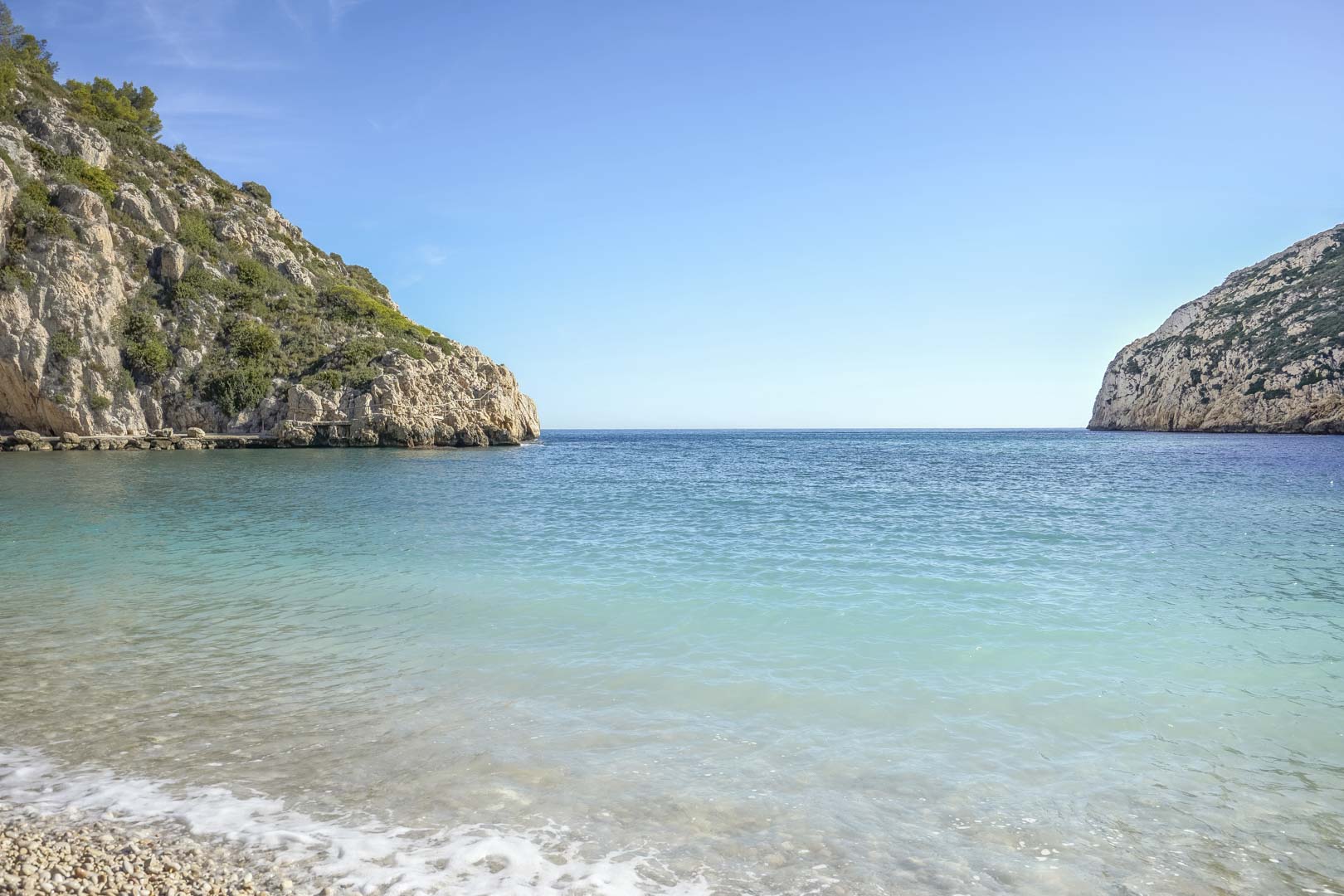 Uitzicht op een mediterrane baai met turquoise water en een kiezelstrand, omgeven door rotsachtige kliffen nabij de woning in Jávea.