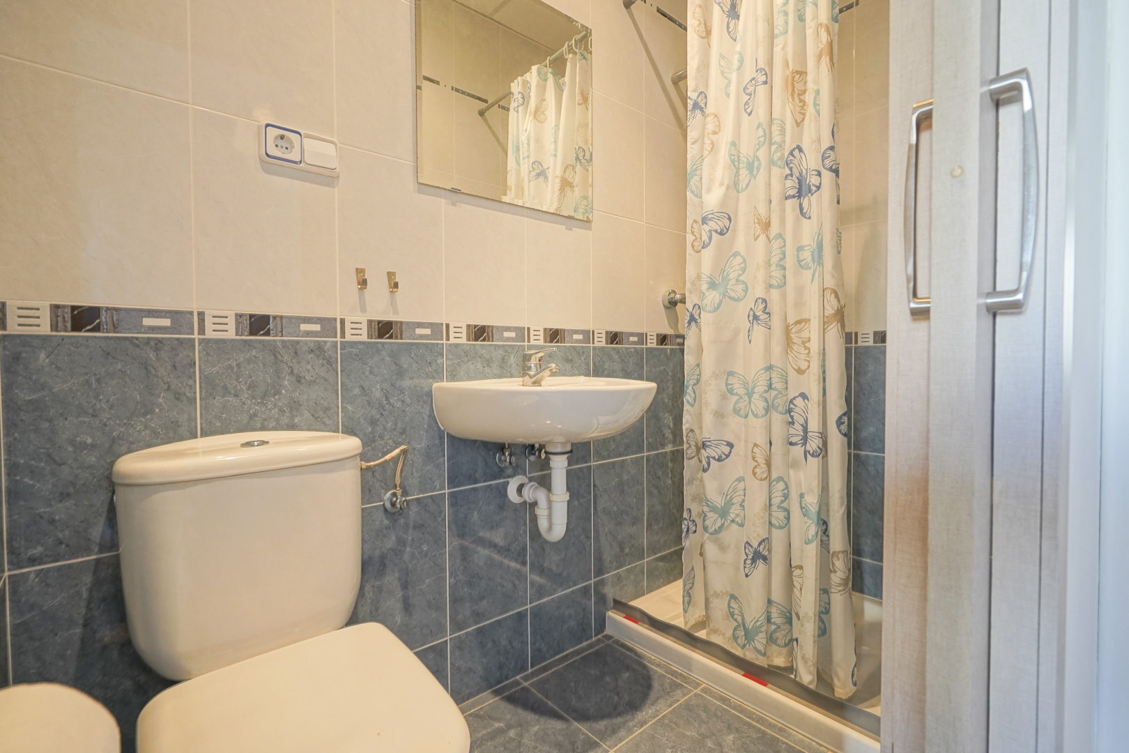 Badkamer met blauwe en crème tegels, voorzien van een wandgemonteerde wastafel, toilet en een doucheruimte met vlindergordijn.