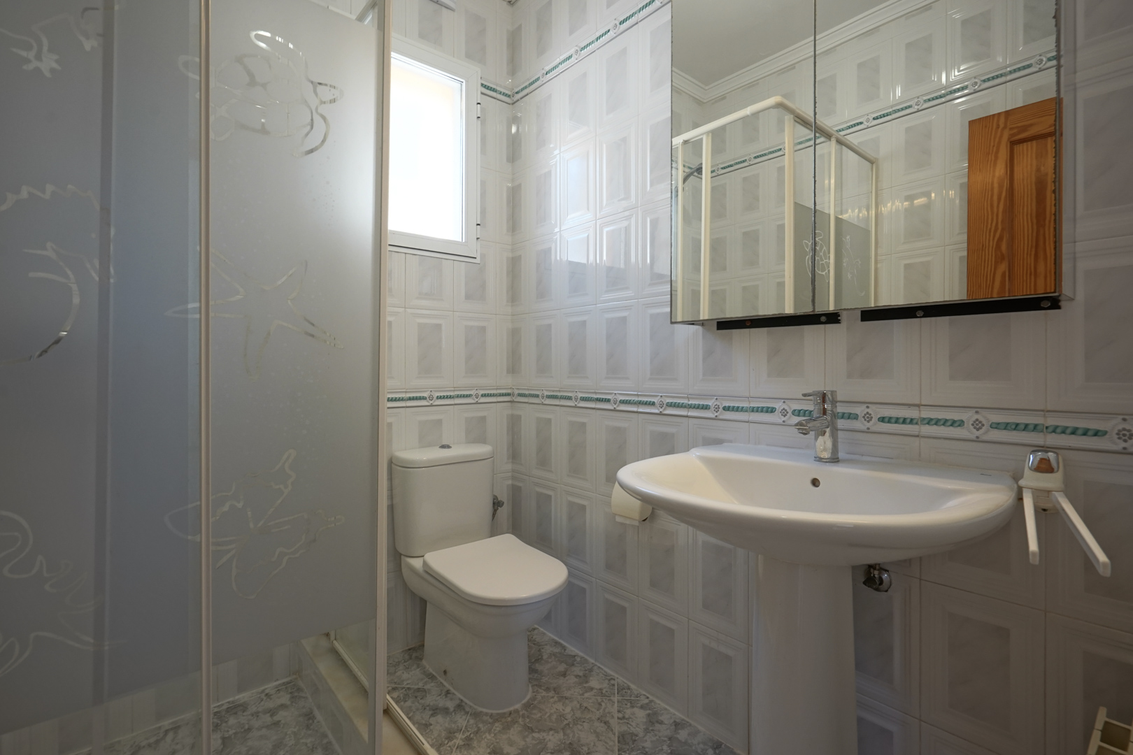 Badkamer voorzien van een wastafel op zuil, toilet en inloopdouche met gezandstraald glas. De wanden zijn afgewerkt met tegels en een decoratieve sierstrip.