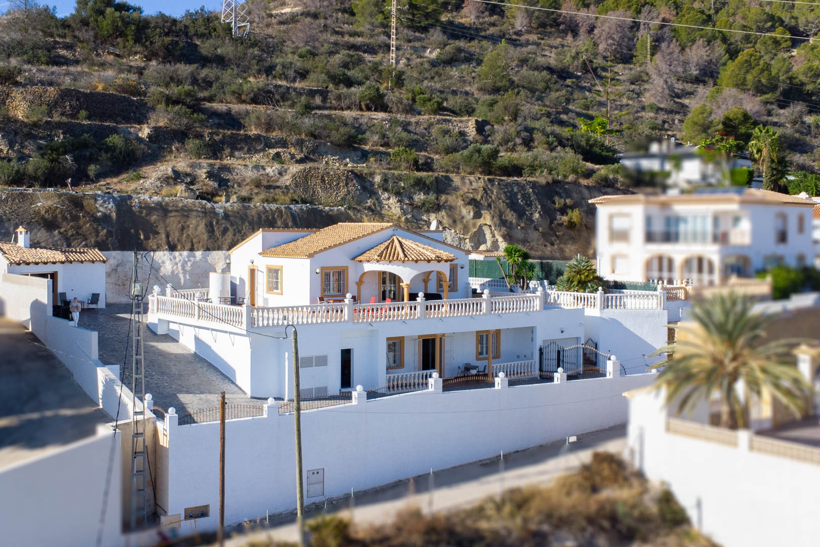 Buitenaanzicht van de villa met twee verdiepingen, witte gevel, terracotta dak en grote terrassen met stenen balustrades aan de voet van de Sierra de Oltà.