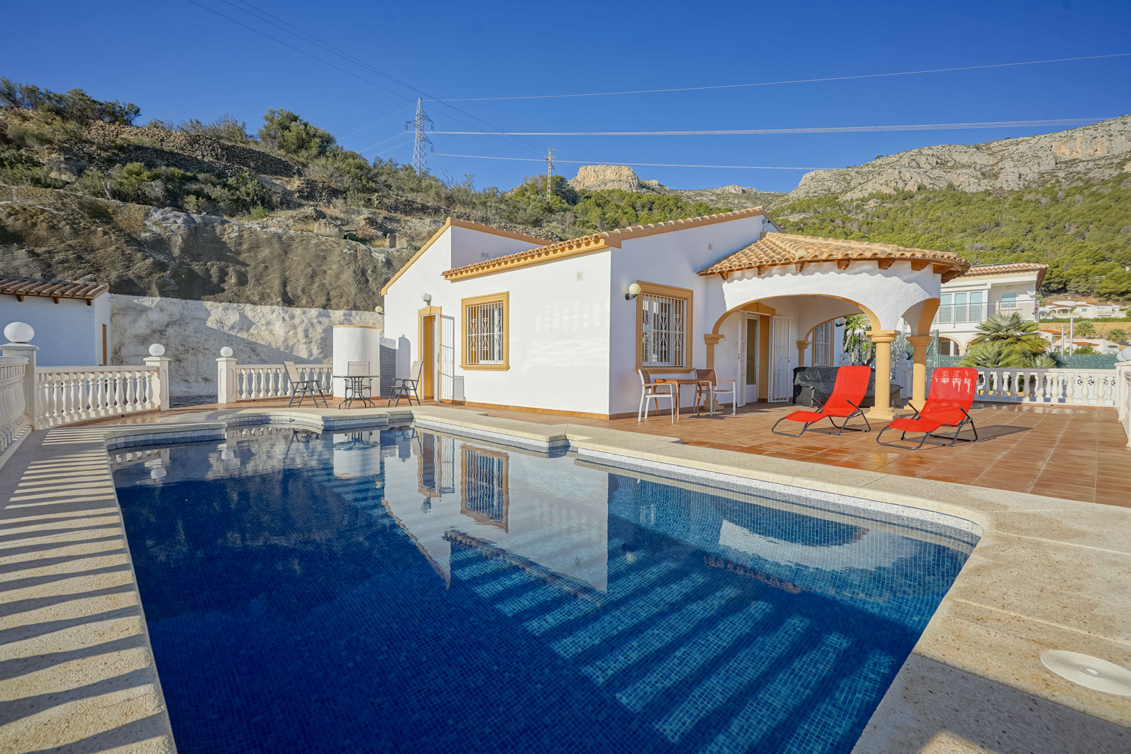 Exterieur van de villa met privézwembad, ruim terras met ligbedden en een overdekte naya met bogen, gelegen aan de voet van de Sierra de Oltà.