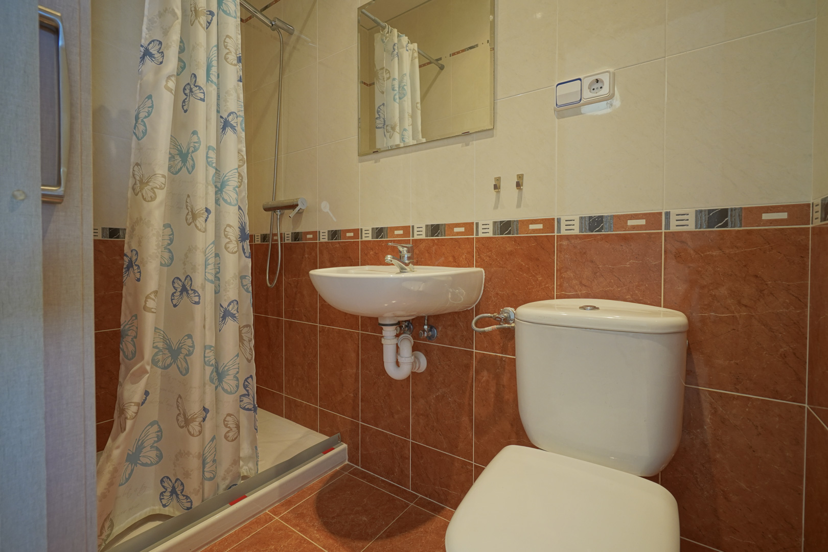 Functionele badkamer met inloopdouche voorzien van vlindergordijn, wandwastafel en tweekleurige betegeling in terra en crème.