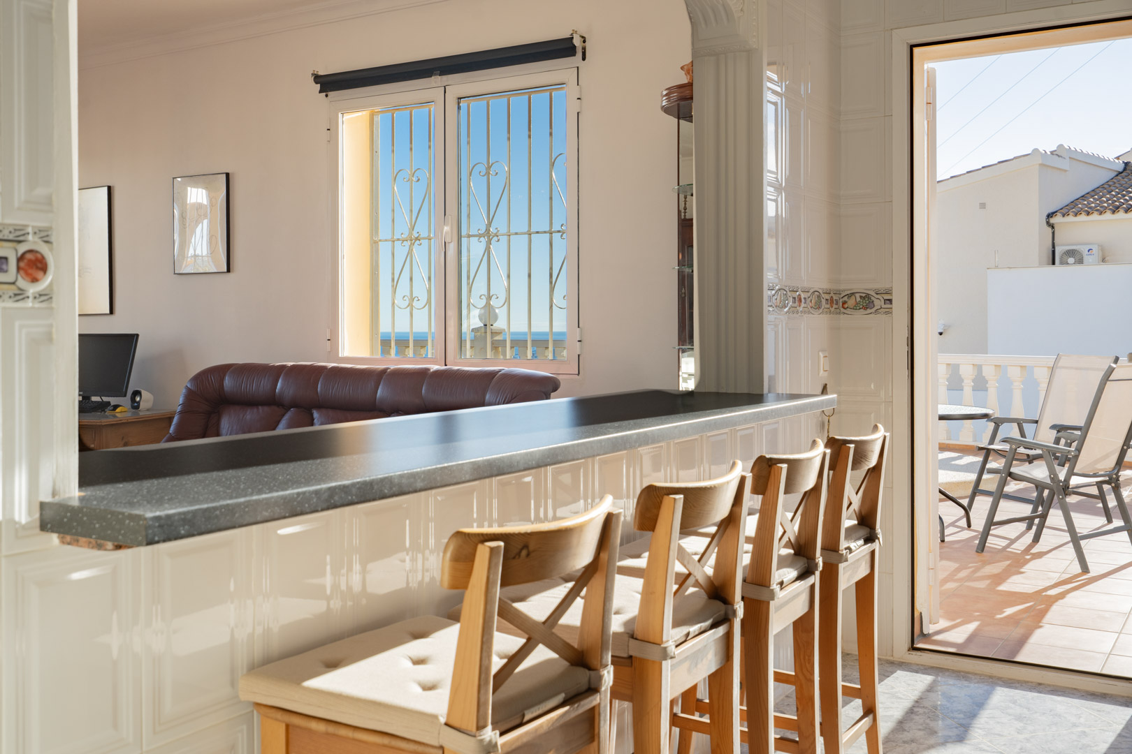 Halfopen keuken met een donkere ontbijtbar en houten krukken, verbonden met de woonkamer en een zonnig terras met uitzicht op zee.