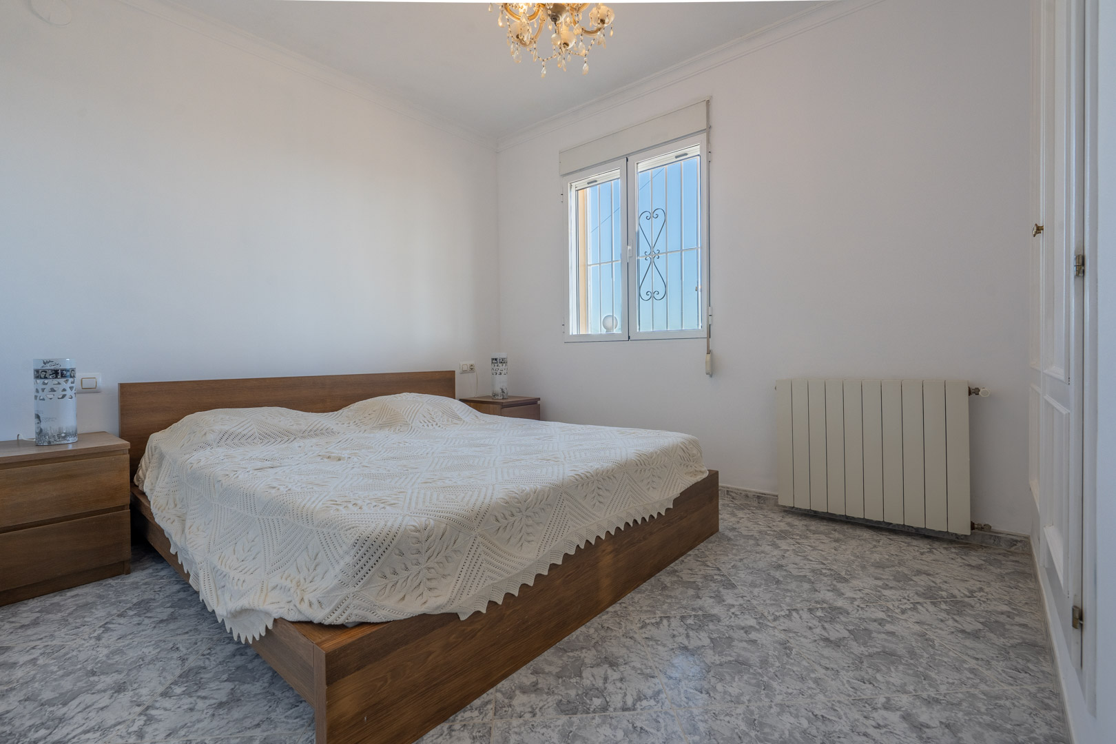 Lichte slaapkamer met tegelvloer in marmerlook, houten bedframe met bijpassende nachtkastjes, witte inbouwkasten en centrale verwarming met radiator.