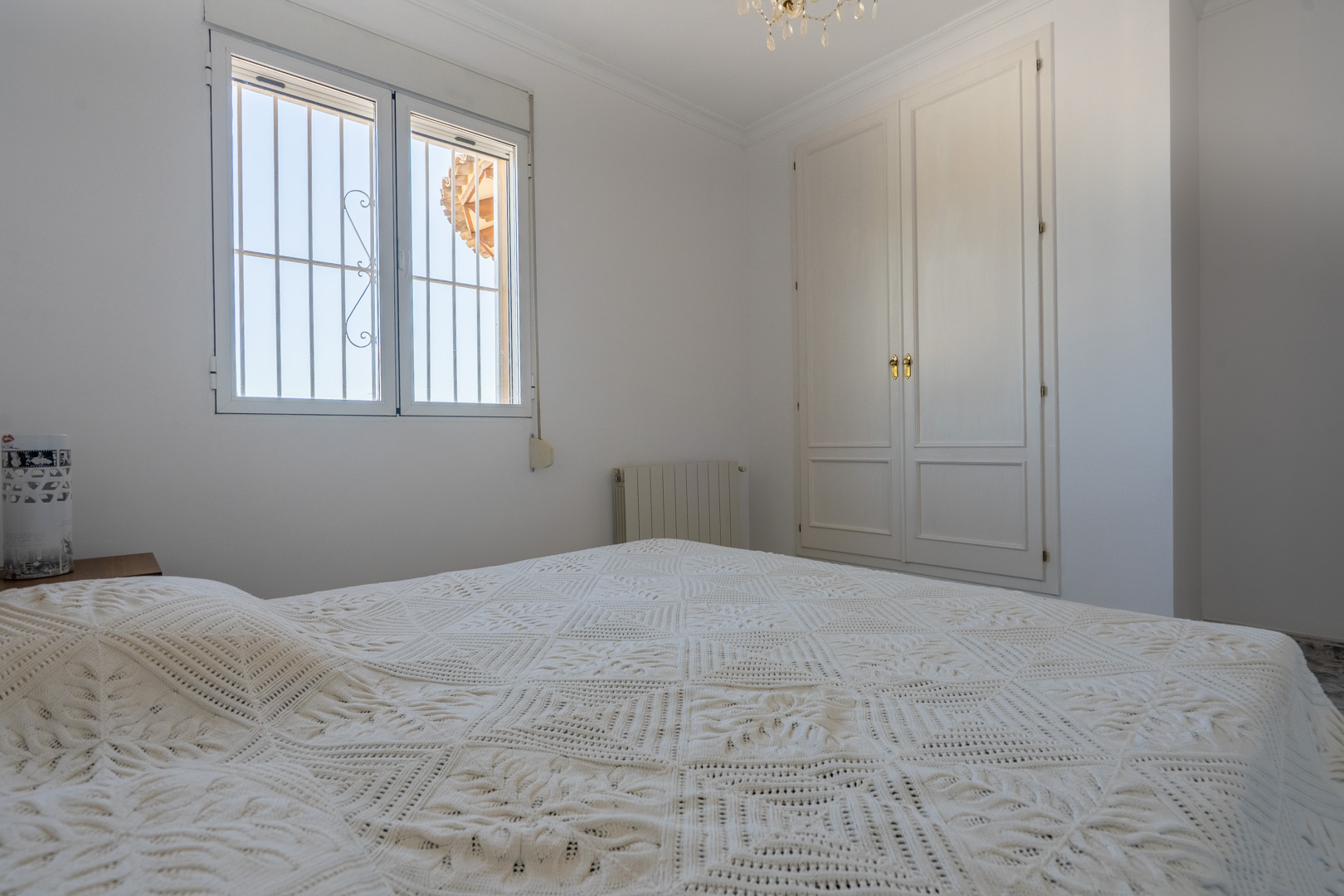 Lichte slaapkamer met witte muren, een ingebouwde houten kledingkast en een raam met beveiligingsroosters. Voorzien van een radiator en veel natuurlijk licht.