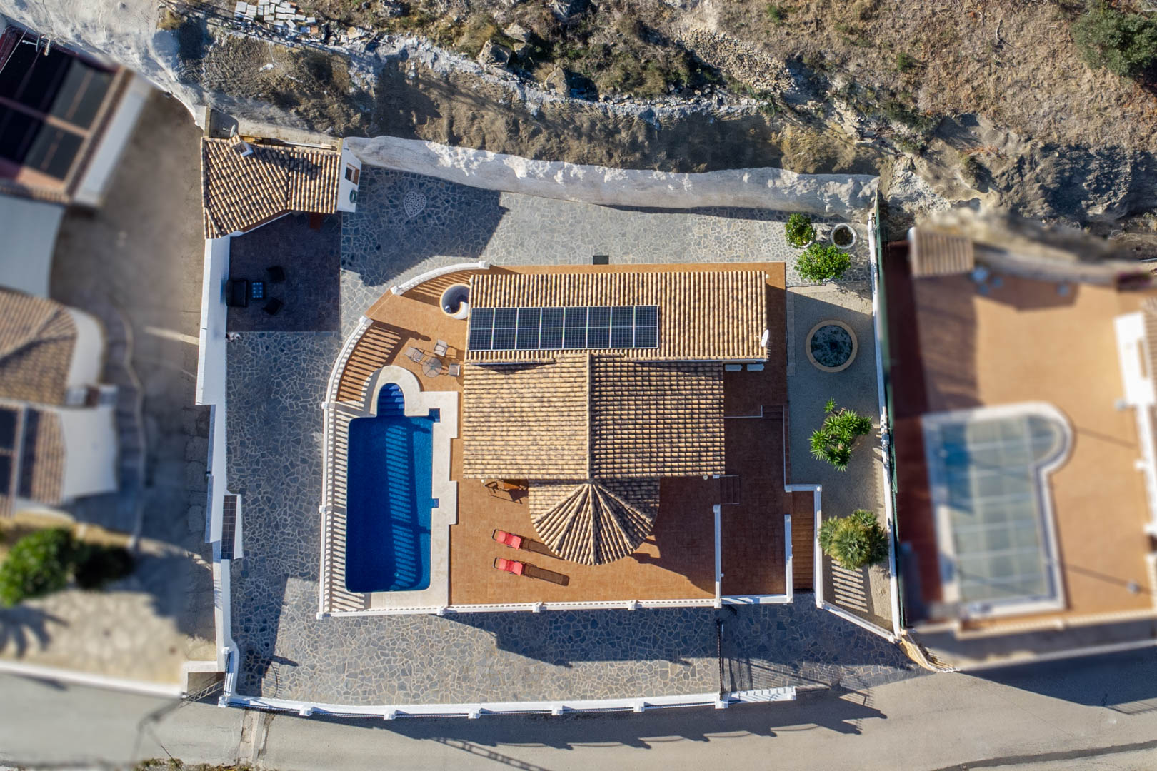 Luchtfoto van de villa met een pannendak voorzien van zonnepanelen, het privézwembad en de diverse betegelde en met steen beklede terrassen.