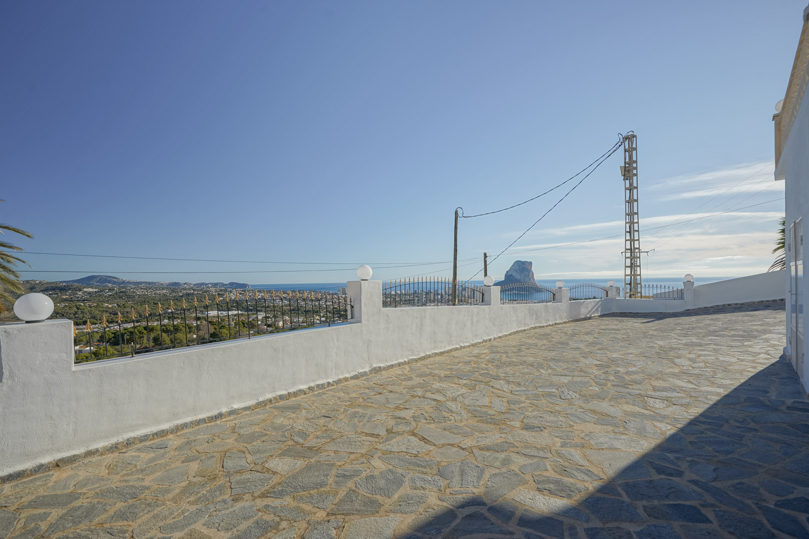 Met natuursteen geplaveid terras met panoramisch uitzicht op de Middellandse Zee en de Peñón de Ifach, omheind door een witte muur met smeedwerk.