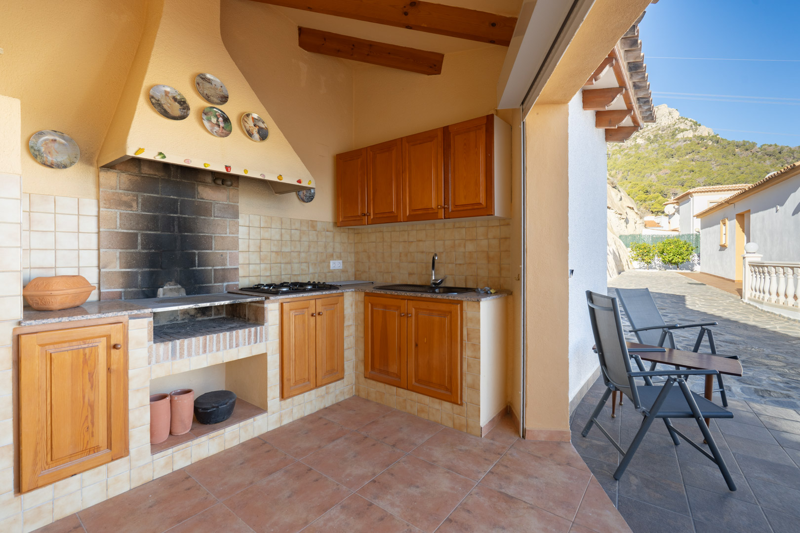 Overdekte buitenkeuken met een gemetselde barbecue, houten kasten, granieten werkblad en spoelbak, grenzend aan een terras met uitzicht op de Sierra de Oltà.