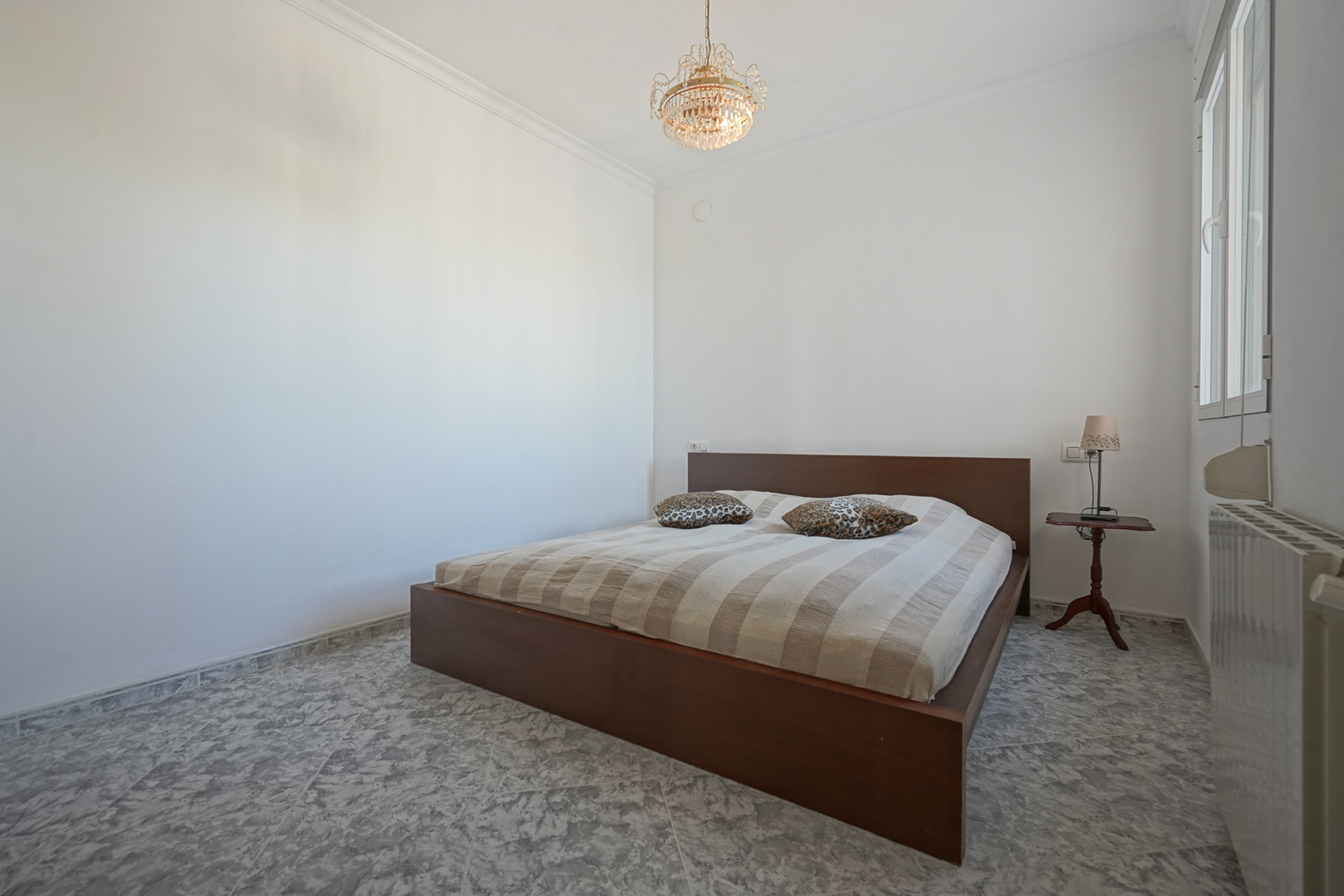Slaapkamer met donkerhouten bedframe, gemarmerde tegelvloer, klassieke kroonluchter en radiator onder het zijraam.