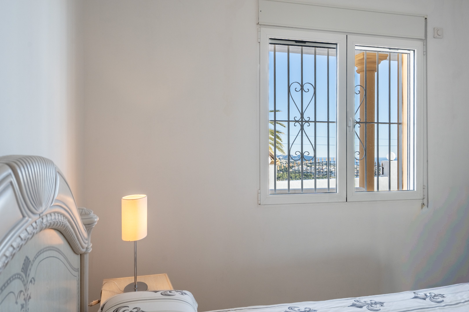 Slaapkamer met klassiek hoofdbord en raam met decoratief traliewerk, met uitzicht op de kustlijn van Calpe en de Middellandse Zee.