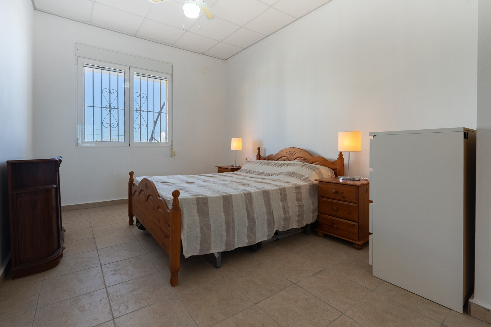 Slaapkamer met traditionele houten meubels, tegelvloer en een raam voorzien van rolluiken en veiligheidstralies voor natuurlijk licht.