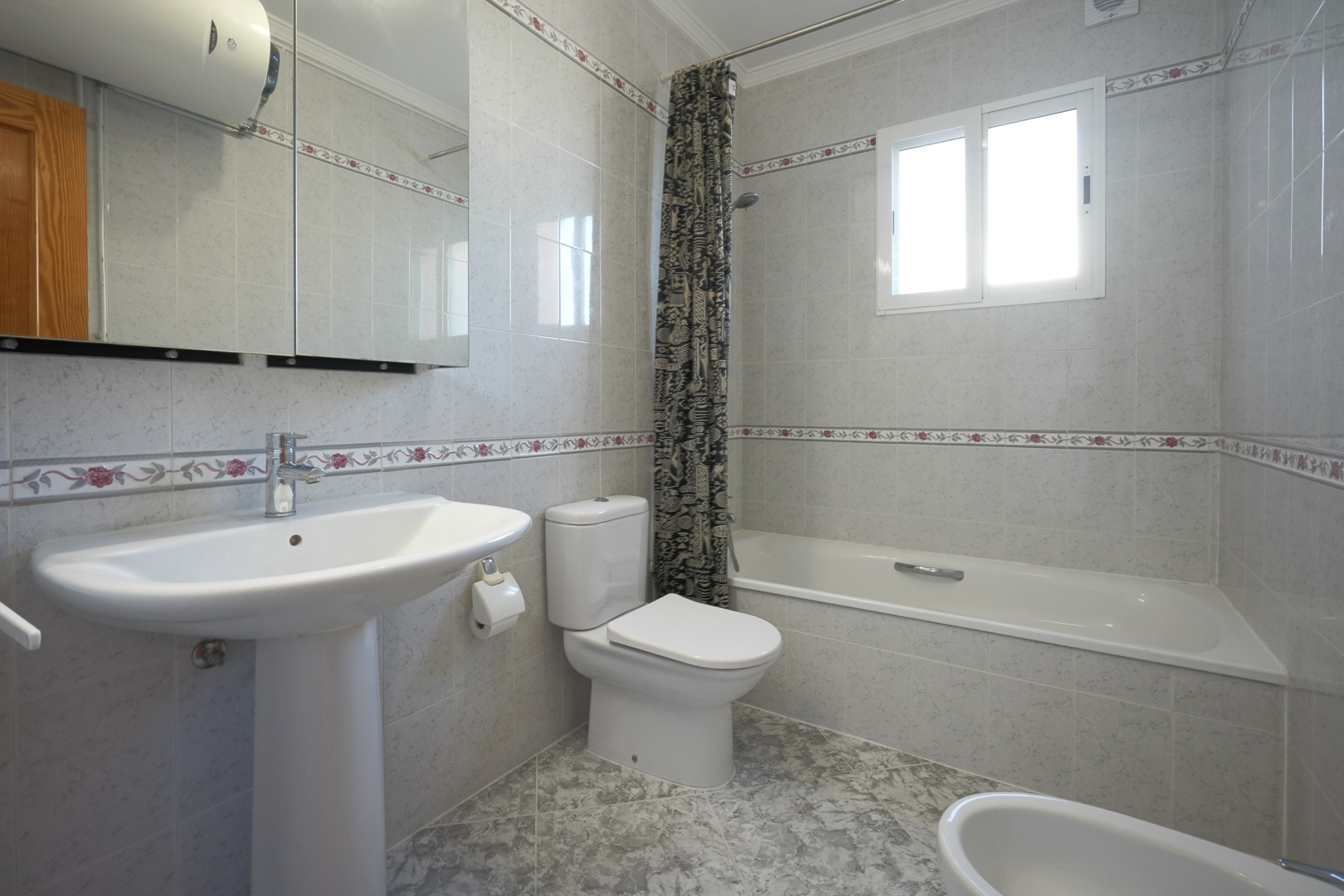 Traditionele badkamer met betegelde wanden en bloemenborders, voorzien van een ligbad, wastafel op zuil en een praktische spiegelkast.