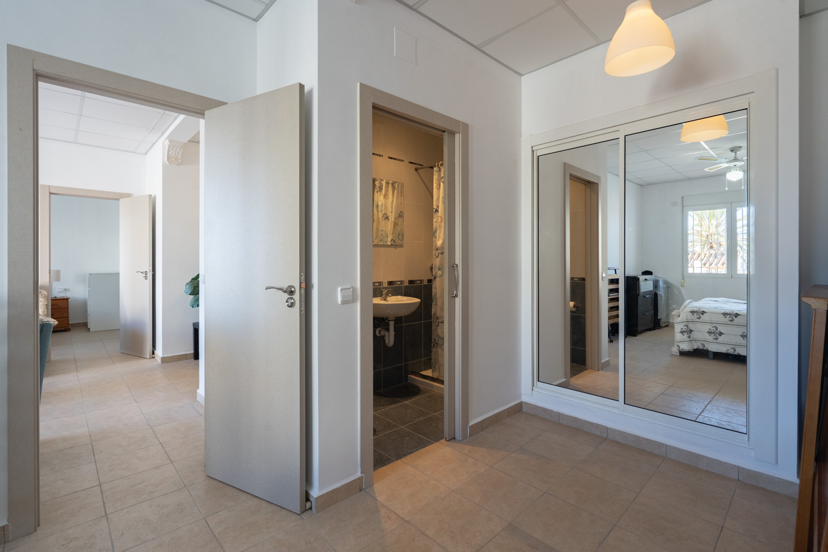 Verdelingsgang met tegelvloer, inbouwkast met spiegeldeuren en toegang tot een badkamer en een slaapkamer voorzien van een plafondventilator.