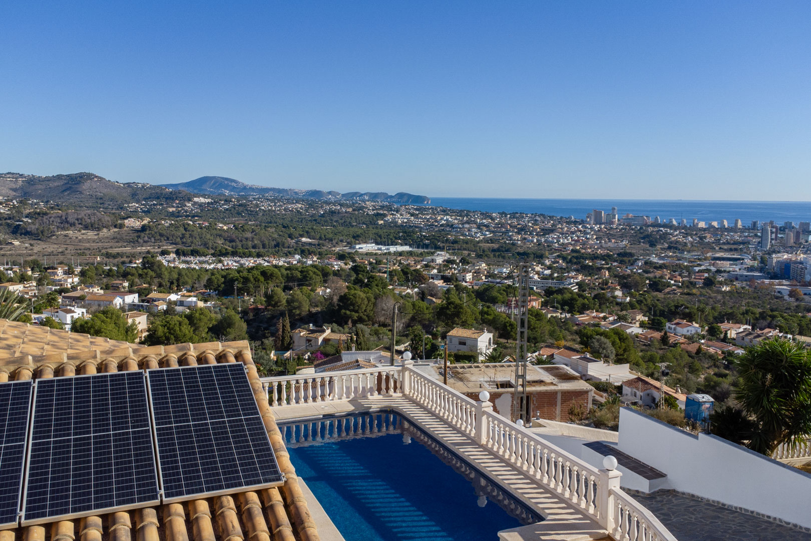 Verhoogd uitzicht op het zwembad met witte balustrades, zonnepanelen op het pannendak en een weids panorama over de kustlijn van Calpe en de Middellandse Zee.
