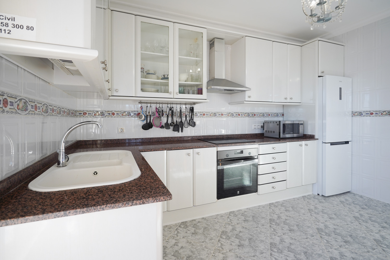 Volledig uitgeruste keuken met witte kasten, roodachtig granieten aanrechtblad en betegelde wanden. Voorzien van inbouwapparatuur en een vloer met marmerlook.