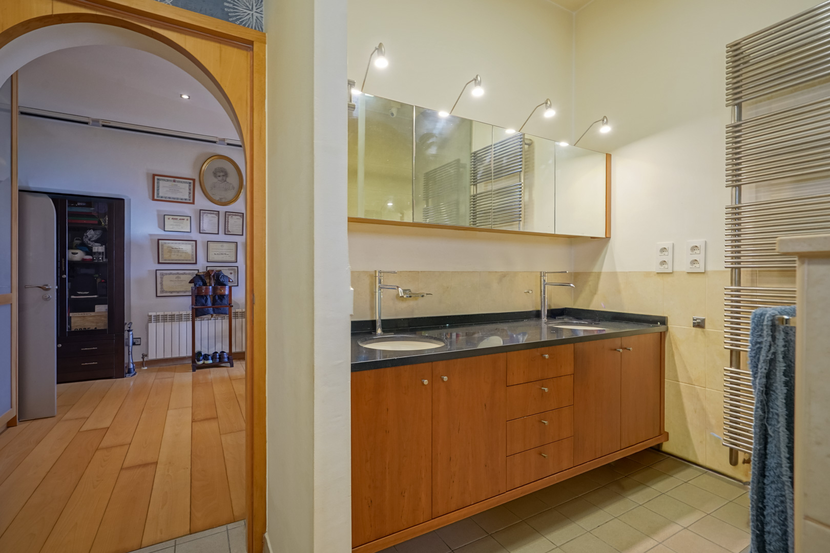 Badkamer met dubbel houten wastafelmeubel, zwart werkblad en verlichte spiegels. Een boogvormige doorgang leidt naar een kleedkamer met hardhouten vloer.