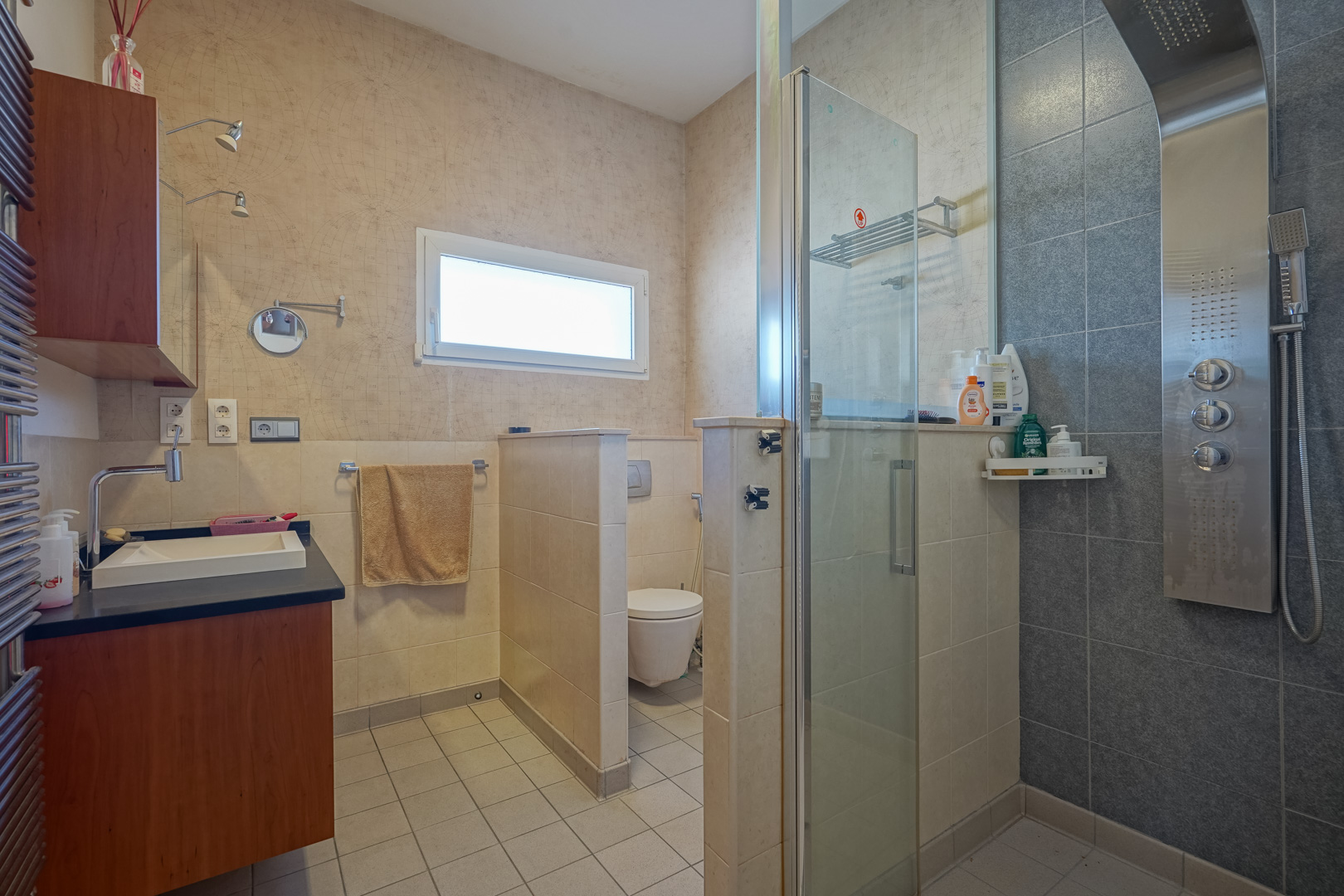 Badkamer met hydromassage douchepaneel, glazen wand en apart toilet. Voorzien van een houten wastafelmeubel met donker blad en behang met wereldkaartmotief.