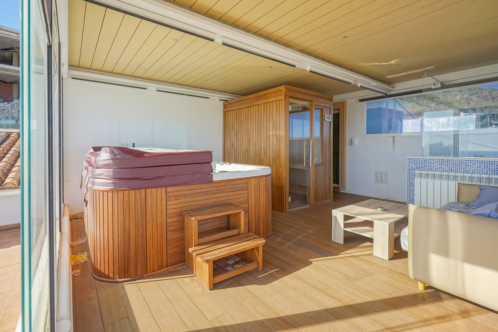 Beglaasd boventerras met wellnessruimte, voorzien van een houten jacuzzi, privésauna en vlonderplanken onder een technisch plafond met geïntegreerde verlichting.
