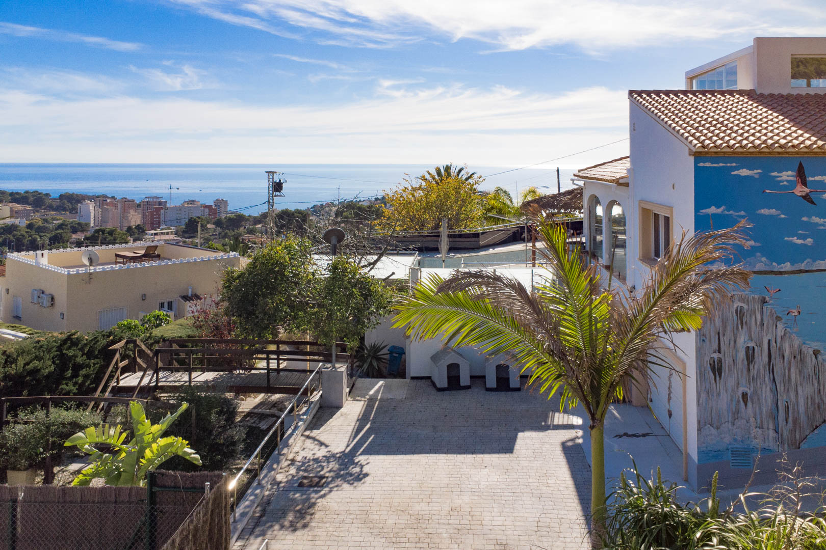 Buitenaanzicht met geplaveide oprit, flamingo-muurschildering op de gevel en panoramisch uitzicht over de Middellandse Zee en de skyline van Calpe.