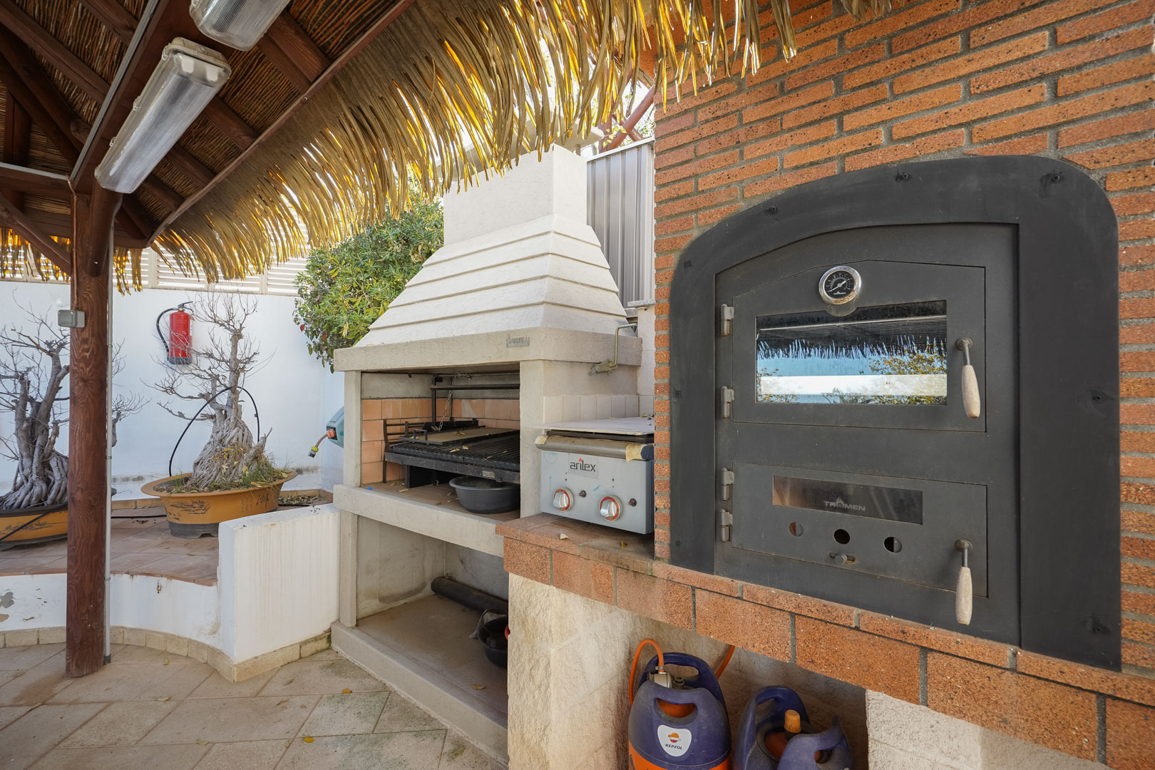 Buitenkeuken voorzien van een ingebouwde Tromen-oven, barbecue en gasgrill onder een rustieke houten pergola met een rieten dak.