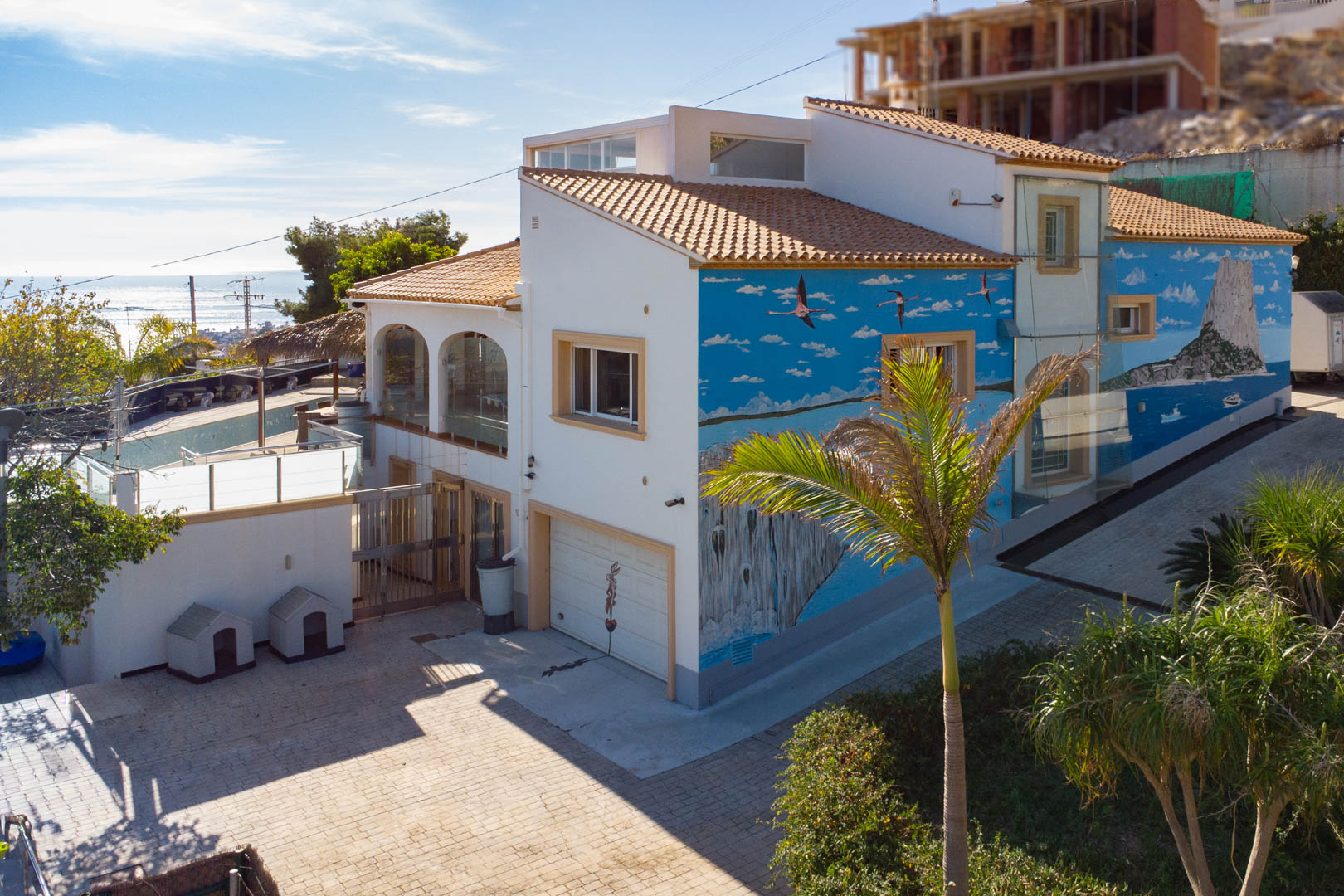 Buitenzicht van de villa met een muurschildering van de Peñón de Ifach, een privégarage en glazen terrassen bij de bestrate oprit.