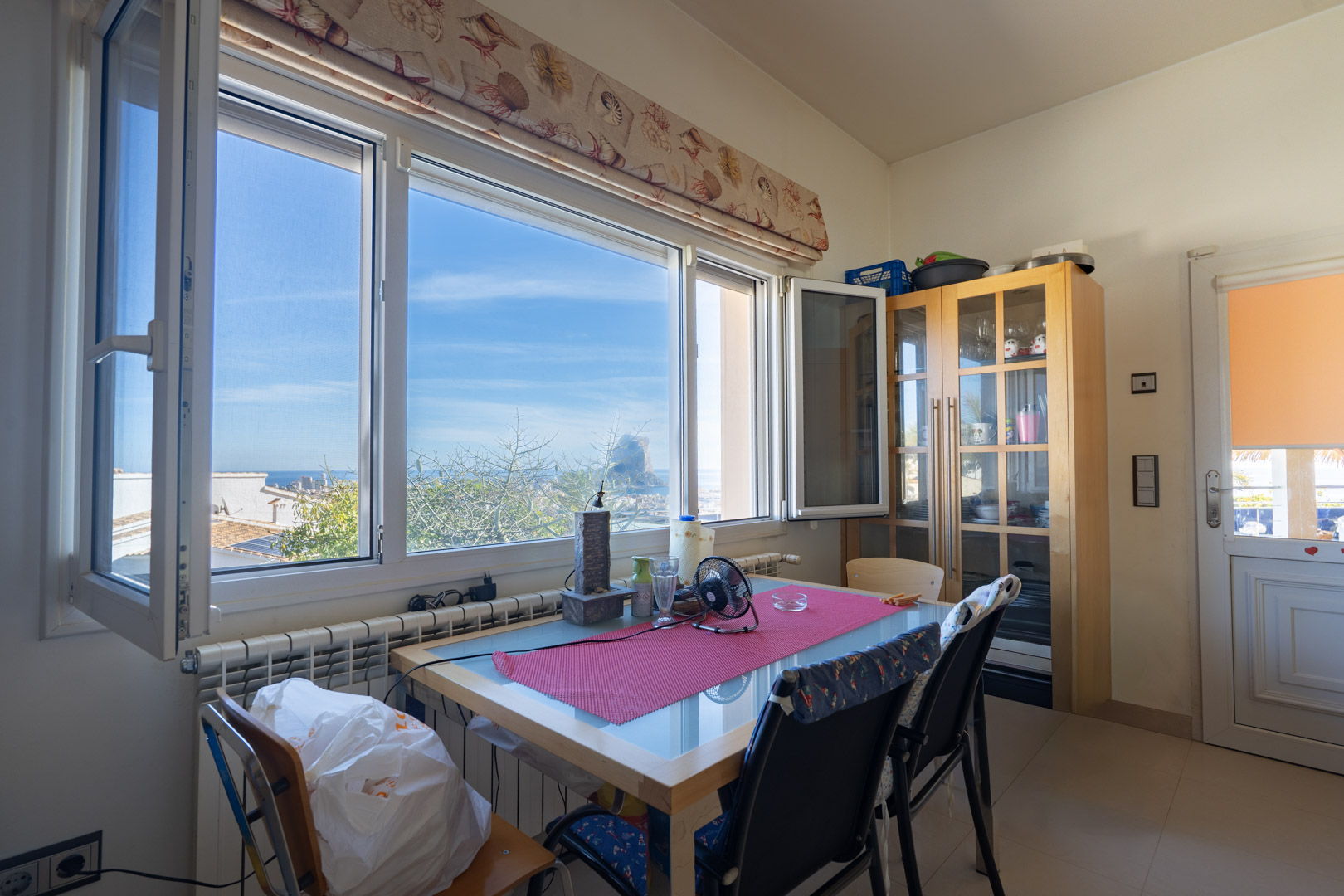 Eethoek met direct uitzicht op de Peñón de Ifach via een groot raam. Voorzien van een houten tafel met glazen blad en een houten vitrinekast.