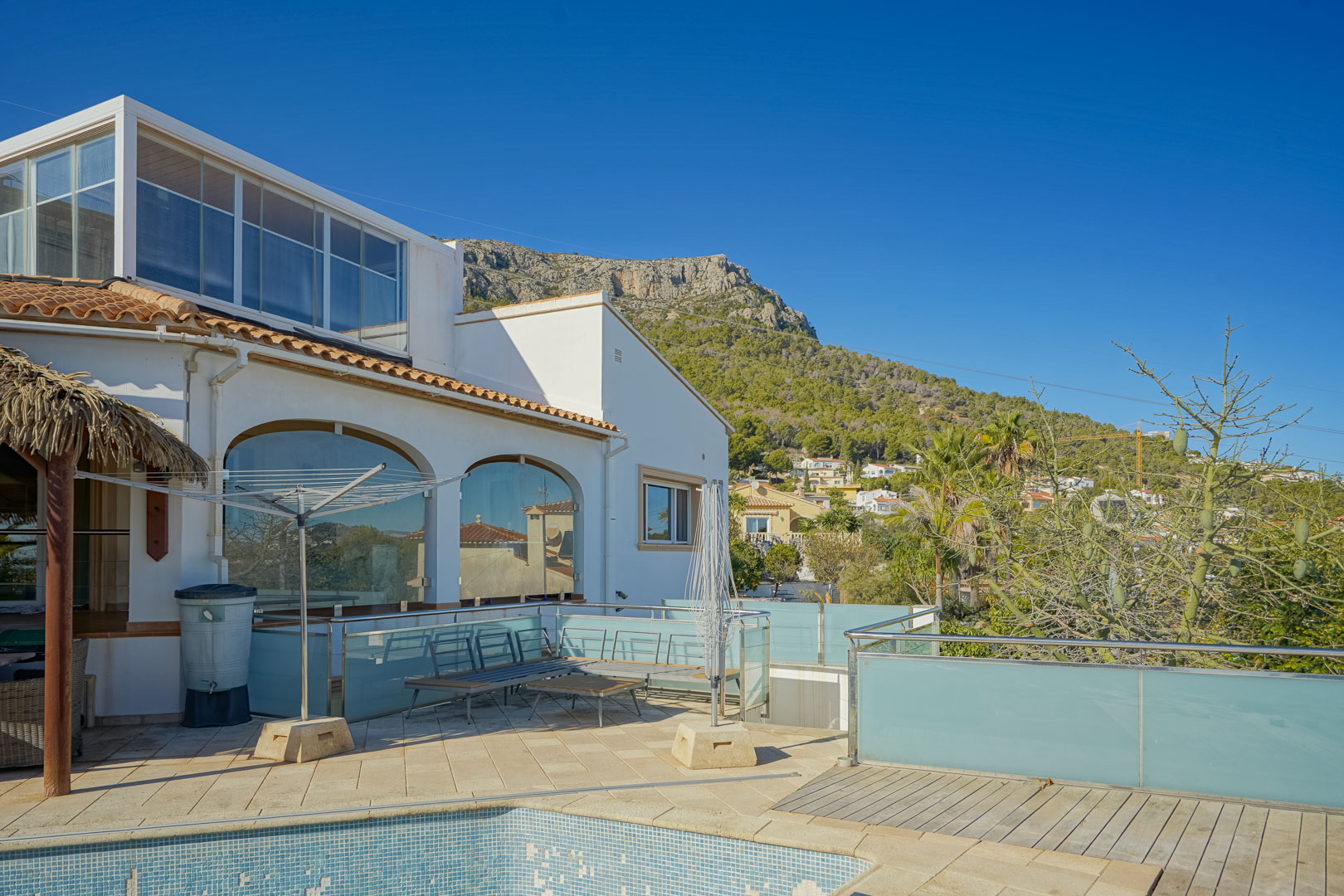 Exterieur van de villa met betegeld terras, glazen balustrades en uitzicht op de Sierra de Oltamar. Voorzien van boogvensters en een glazen bovenbouw bij het zwembad.