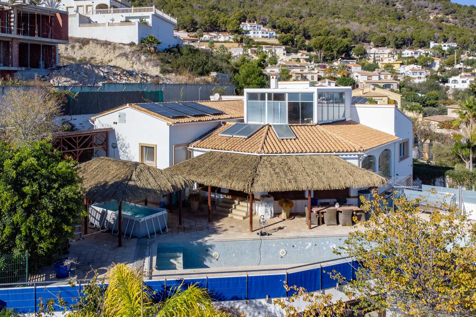 Exterieur van de villa met zonnepanelen, terrassen met rieten pergola's, zwembad en een glazen opbouw op het bovenste niveau in de heuvels van Calpe.
