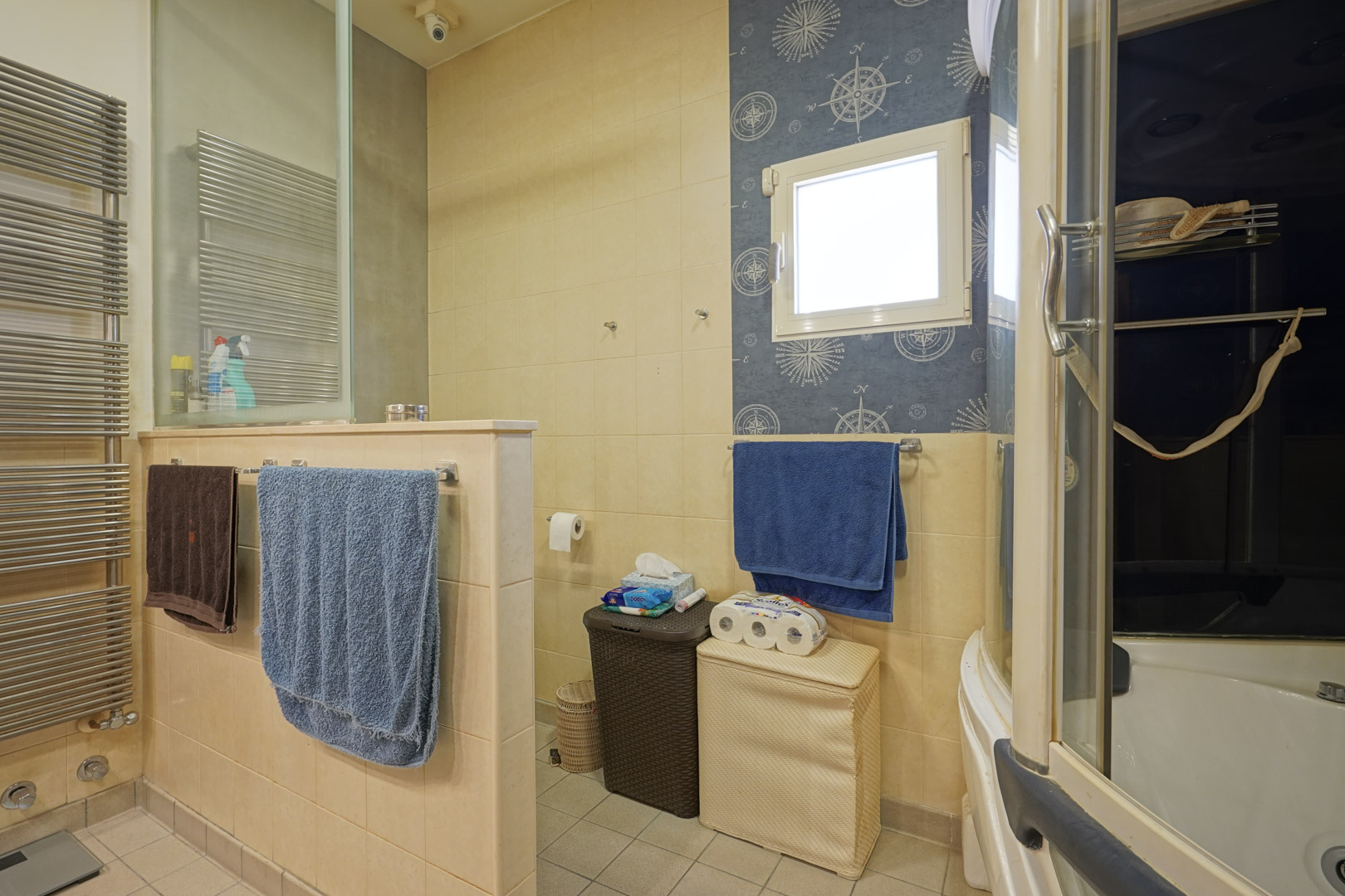 Functionele badkamer met crème keramische tegels, een design handdoekradiator en een afgescheiden toiletruimte. De wand bij het raam is voorzien van nautisch behang.