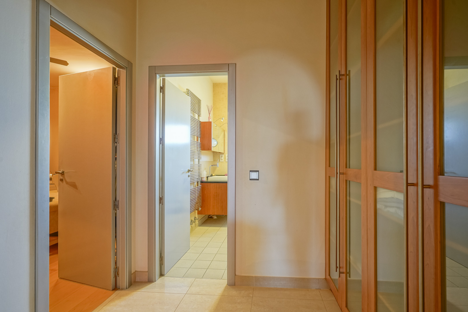Functionele hal met ingebouwde houten kasten met glaspanelen, die een slaapkamer met hardhouten vloer verbindt met een en-suite badkamer met handdoekradiator.