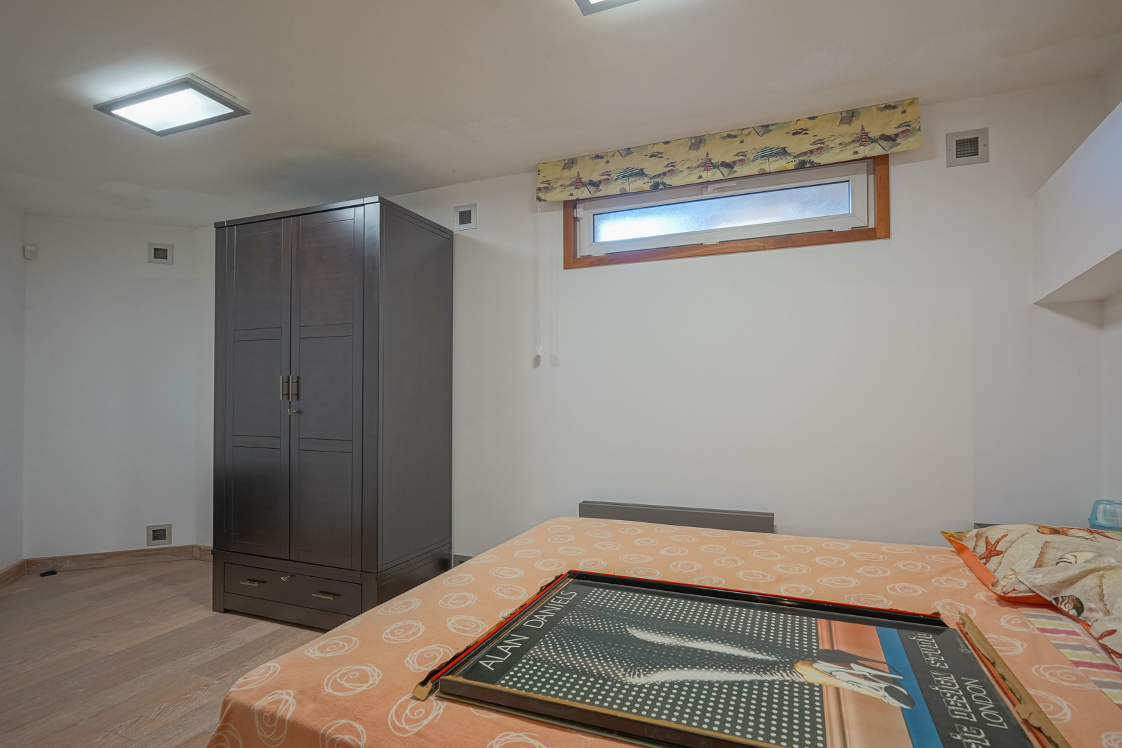 Functionele slaapkamer met een donkerhouten kledingkast, lichte laminaatvloer en een horizontaal raam onder vierkante inbouwspots in het plafond.