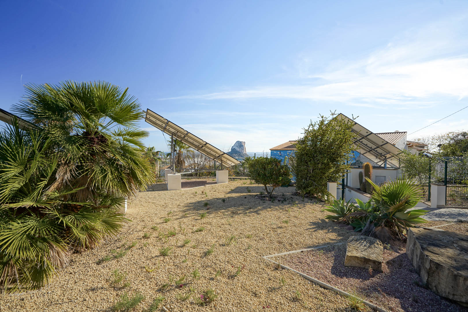 Geavanceerd fotovoltaïsch systeem met gemotoriseerde zonnetorens in een grindtuin, met uitzicht op de Peñón de Ifach en mediterrane planten.