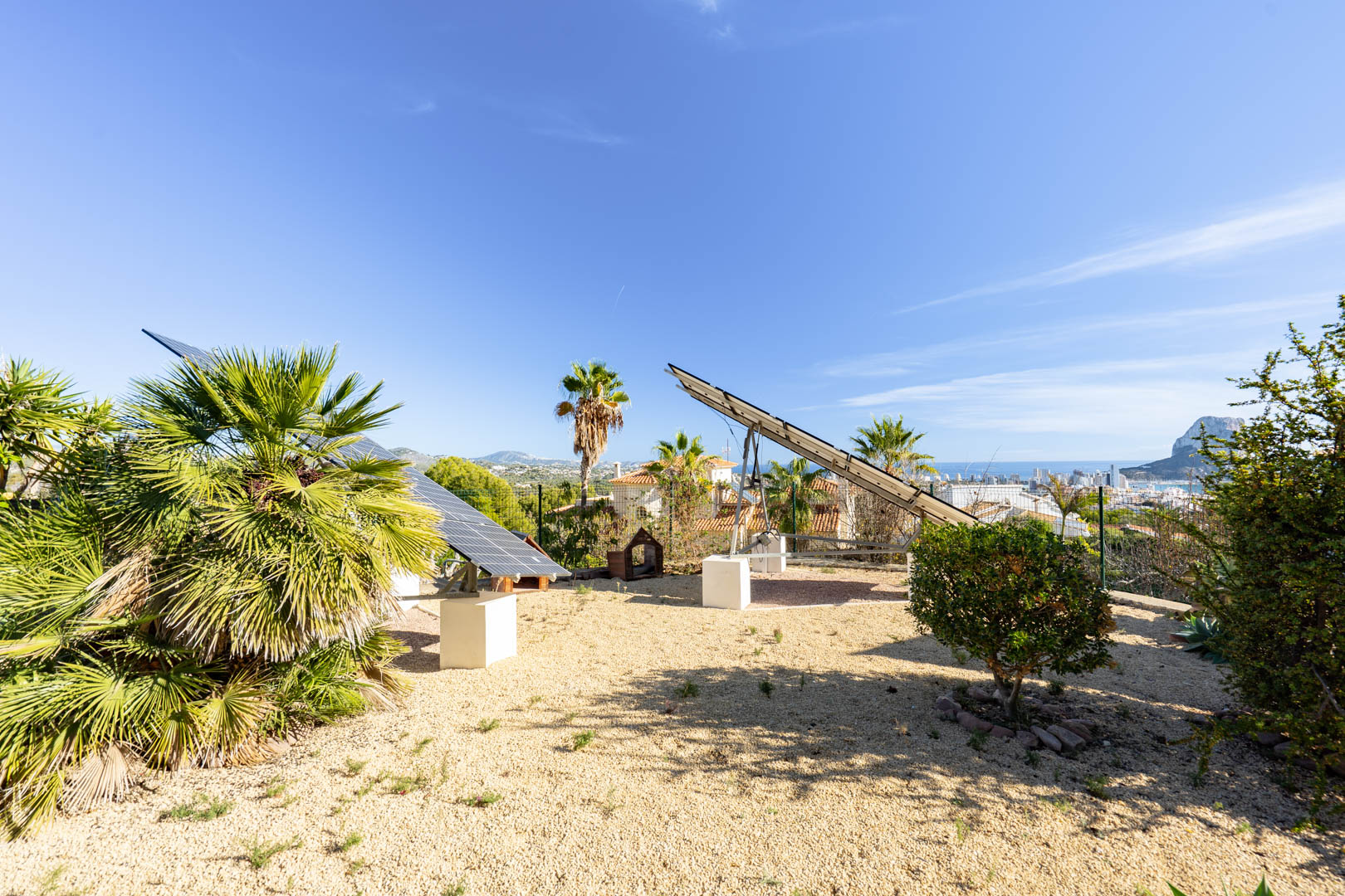 Gemotoriseerde zonnepanelen op betonnen sokkels in een grindtuin, met panoramisch uitzicht op de Peñón de Ifach en de skyline van Calpe.