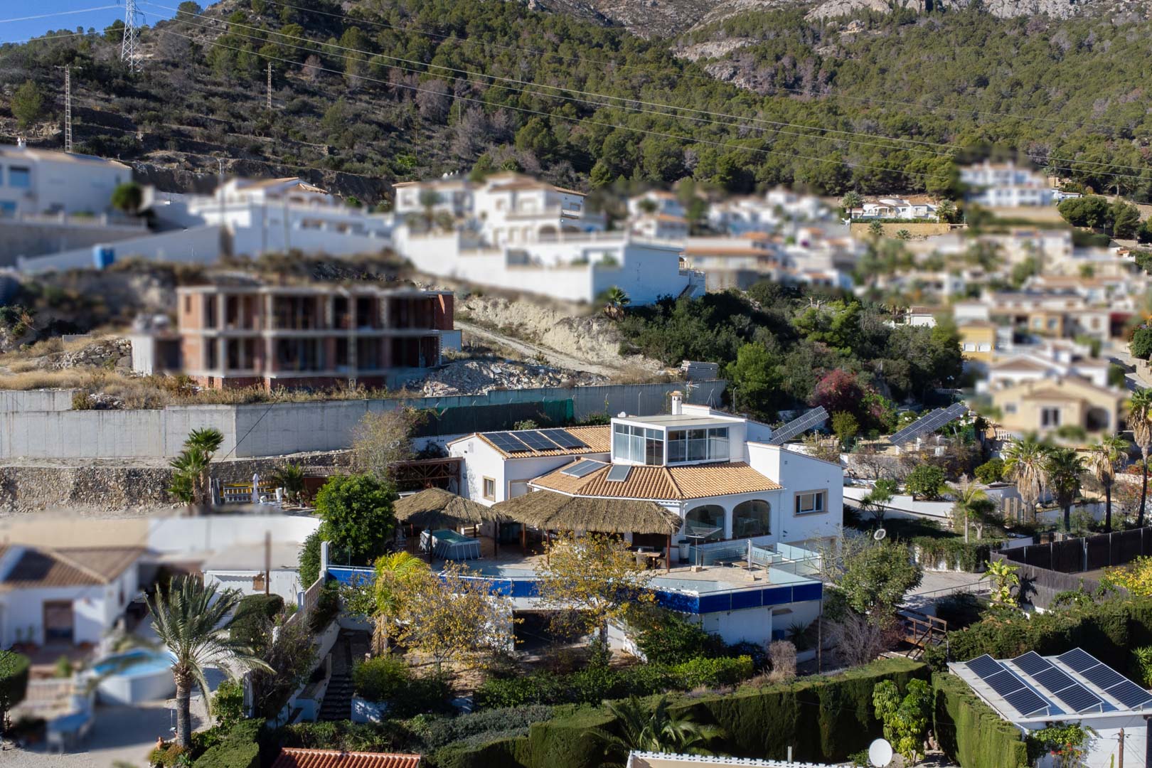 Gevelzicht van de villa met meerdere niveaus, zonnepanelen, een glazen bovenverdieping en ruime terrassen met rieten pergola's, gelegen tegen de helling in Calpe.